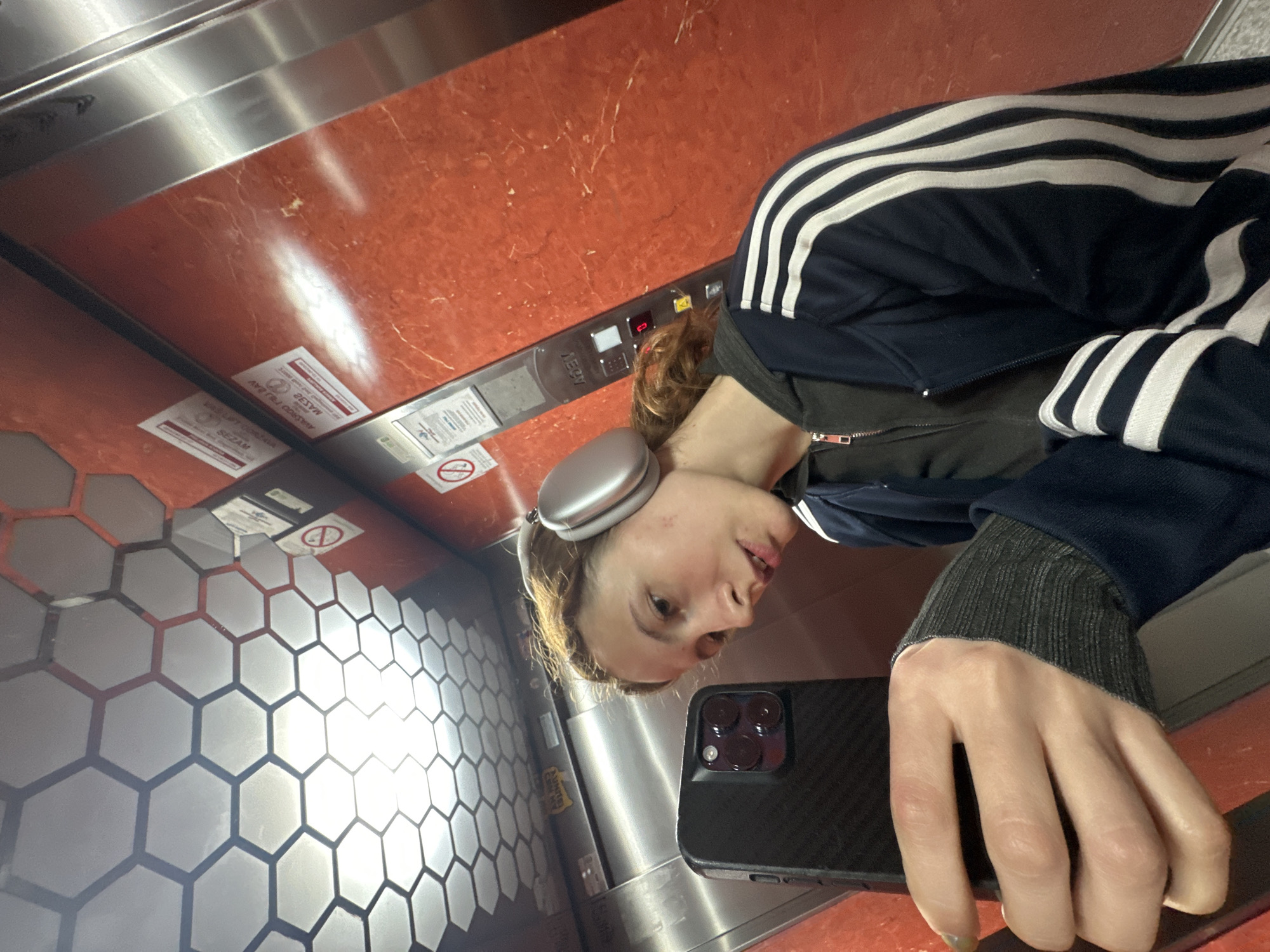 The image is a selfie taken by a young person inside an elevator, originally rotated 90 degrees counter-clockwise. The subject, a young individual with light brown hair and silver over-ear headphones, is wearing a navy blue tracksuit jacket with white stripes on the sleeves, layered over a dark long-sleeved top with ribbed cuffs. They are holding a black smartphone with a textured case and a multi-lens camera setup, looking towards the lens with a neutral expression.
The elevator interior features a combination of materials. Some wall panels are polished metallic, reflecting light, while others are a reddish-brown, possibly marble, material with lighter veining. A distinctive section of the wall is decorated with reflective hexagonal tiles, forming a honeycomb pattern. An elevator control panel is visible, displaying the number "2" and a downward arrow, along with various buttons.
Several inverted signs are affixed to the elevator walls. One red and white sign clearly states "MASKE SU OBAVEZNE" (Masks are mandatory) and "POŠTUJTE FIZIČKU DISTANCU" (Respect physical distance), indicating adherence to public health guidelines. Another inverted sign partially visible reads "SEZAM." A universal prohibition symbol (red circle with a diagonal line) is also present on one of the signs. The scene is illuminated by artificial indoor lighting, casting reflections on the metallic surfaces. The location context provided is Novi Sad, Serbia.