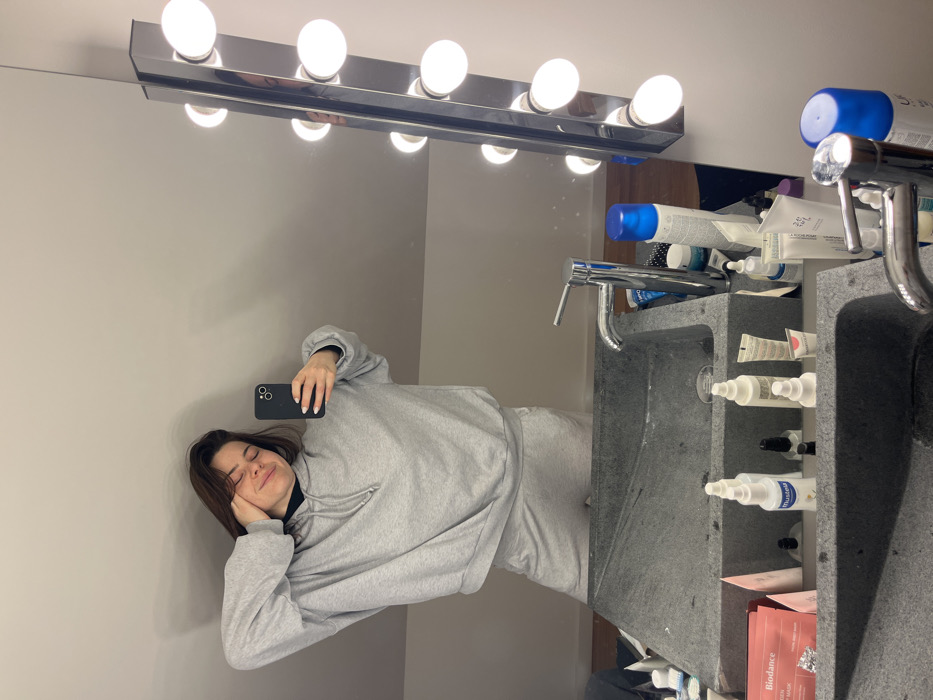 A young woman is captured in a mirror selfie, reflecting a relaxed moment in what appears to be a modern bathroom. The image is oriented sideways, with the woman positioned on the left side of the frame as if she were standing upright.
The woman has shoulder-length brown hair and is wearing a light gray oversized hooded sweatshirt and matching sweatpants, suggesting casual and comfortable attire. Her eyes are closed, and a gentle, contented smile plays on her lips, conveying a sense of peace or drowsiness. She holds a black smartphone in her left hand, extending it to take the selfie, while her right hand is casually resting near her jaw.
Above the mirror, a chrome or silver-toned vanity light fixture spans horizontally, fitted with five prominent spherical white bulbs that are illuminated, casting bright light on the scene. The wall reflected behind the woman is a plain, light gray, indicating a simple background.
To the right of the woman, the reflection reveals a contemporary bathroom vanity area. The counter and sinks appear to be constructed from a dark gray, textured material, possibly concrete or a similar stone, featuring clean, angular lines. At least two sinks are visible, suggesting a double vanity setup. The sink closer to the woman (on the left side of the vanity) has a sleek, single-lever chrome faucet.
The vanity surface and integrated shelves are adorned with a collection of toiletries and beauty products. Various bottles, tubes, and containers, many with blue or white caps, are scattered around the sinks. Several specific brand names are visible: "BIODANCE" can be seen on a reddish-pink packaged item, likely a face mask, and "MUSTELA" is discernable on a white bottle with a blue cap. Other white tubes and bottles, possibly skincare items, are also present, some appearing to be from brands like La Roche-Posay, though the text is not entirely clear.
The overall impression is one of a candid, quiet moment, perhaps captured early in the morning or late at night, in a well-lit and organized, yet lived-in, bathroom setting.