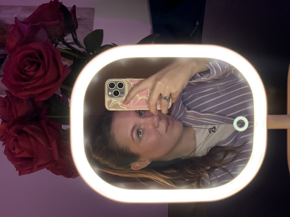 The image displays a mirror selfie taken indoors in Belgrade, Serbia. A Caucasian female is reflected in an illuminated, oval-shaped vanity mirror with an integrated LED light strip and a circular touch-sensitive power button at its base. She holds an iPhone in her right hand, partially obscuring the right side of her face. The phone case features a light background with pink floral motifs. She wears a light purple and white striped shirt over a white undershirt, a dark, abstract-shaped pendant necklace, and a ring with a dark faceted stone on her right ring finger. Her light brown hair is pulled into a ponytail, and a small stud earring is visible in her left ear. She looks directly towards the phone's camera, with a slight, neutral expression. In the background, behind the mirror, a bouquet of deep red roses is visible against a wall illuminated by a reddish-purple ambient light. A dark, smooth surface, likely a tabletop, is present beneath the mirror.
