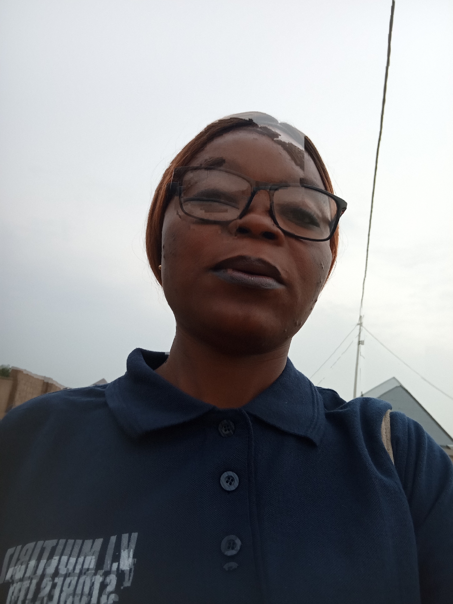A woman, possibly in her late 20s to early 30s, is captured in a close-up, low-angle selfie. She has dark skin, brown hair styled back, and is wearing dark-rimmed glasses. Her lips are painted with dark lipstick. She is wearing a dark blue polo shirt with buttons at the collar. A light-colored strap, possibly from a bag, is visible over her right shoulder. In the background, the sky is overcast, suggesting a cloudy day. To the left, a section of a building with a rough texture is visible. To the right, a thin wire extends upwards, disappearing into the sky, and a portion of a building with a triangular roof can be seen. The overall environment suggests an outdoor urban setting, possibly in Kason, Nigeria. The focus of the image is on the woman's face, with the background slightly blurred. There is text visible on the woman's shirt, but it is partially obscured and not legible.