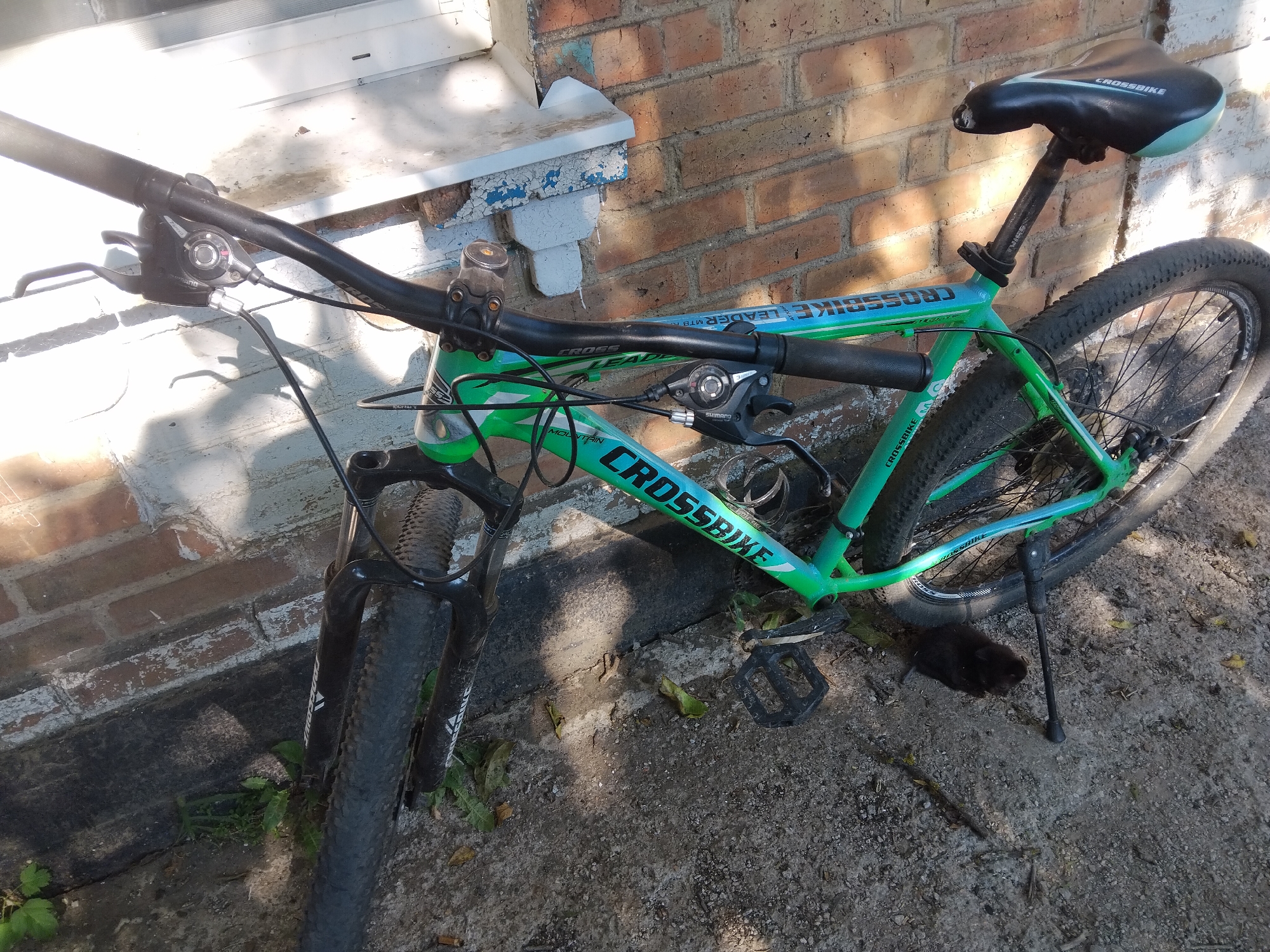 An outdoor, medium-angle shot shows a green and black mountain bicycle leaning against a brick wall in Pervozvanivka, Ukraine. The bicycle is upright, supported by a kickstand. Its frame is predominantly green with blue accents and the brand "CROSSBIKE" is visible in white lettering. The handlebars are black, with brake levers and a gear shifter. The bicycle has a black seat with white "CROSSBIKE" branding and a suspension fork. The front wheel is turned slightly to the left, and the rear wheel is visible. The ground beneath the bicycle is a mixture of dirt, gravel, and some dried leaves. The brick wall behind the bicycle is weathered, with patches of peeling paint on a white ledge above it. Sunlight casts shadows across the scene, indicating it is daytime. No people are present.
