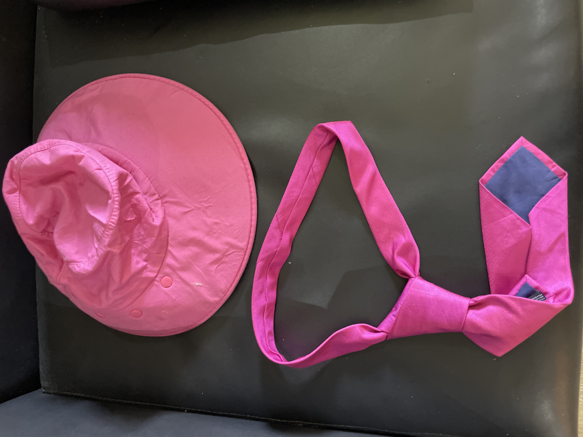 In Ado, Nigeria, on a black leather surface, a bright pink sun hat and a matching pink necktie lie. The sun hat is circular with a wide brim and a gathered crown, featuring two pink buttons on the side. The necktie is knotted and laid out in a U-shape, revealing a dark blue lining at its tip. The lighting is artificial, creating soft shadows.