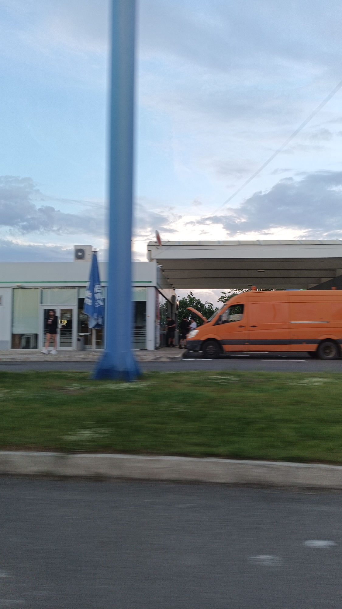 The image depicts a gas station on the side of a road. An orange van is parked under the gas station canopy with its hood open. Several people are standing near the van. A person is seen walking near the convenience store portion of the gas station. A blue pole is in the foreground, slightly obscuring the view. The sky is overcast with clouds. The location appears to be in Burgas, Bulgaria.