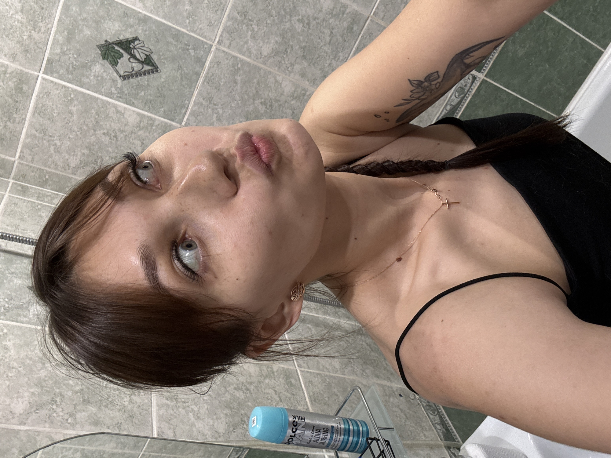 A young woman with brown hair, parted bangs, and light blue eyes is looking upwards and to the right with her lips pursed. She is wearing a black spaghetti strap top and a thin gold necklace with a cross pendant. Her right arm is visible, showing a tattoo of a flower and bird. She has a single gold earring in her left ear. To her left, a can of "DOLCE MAN MILK Fresh & Citrus Aloha Surfing" product is on a shelf. The background consists of green and white tiled walls, typical of a bathroom. There is no information available to suggest this image is from Novokuznetsk, Russia.