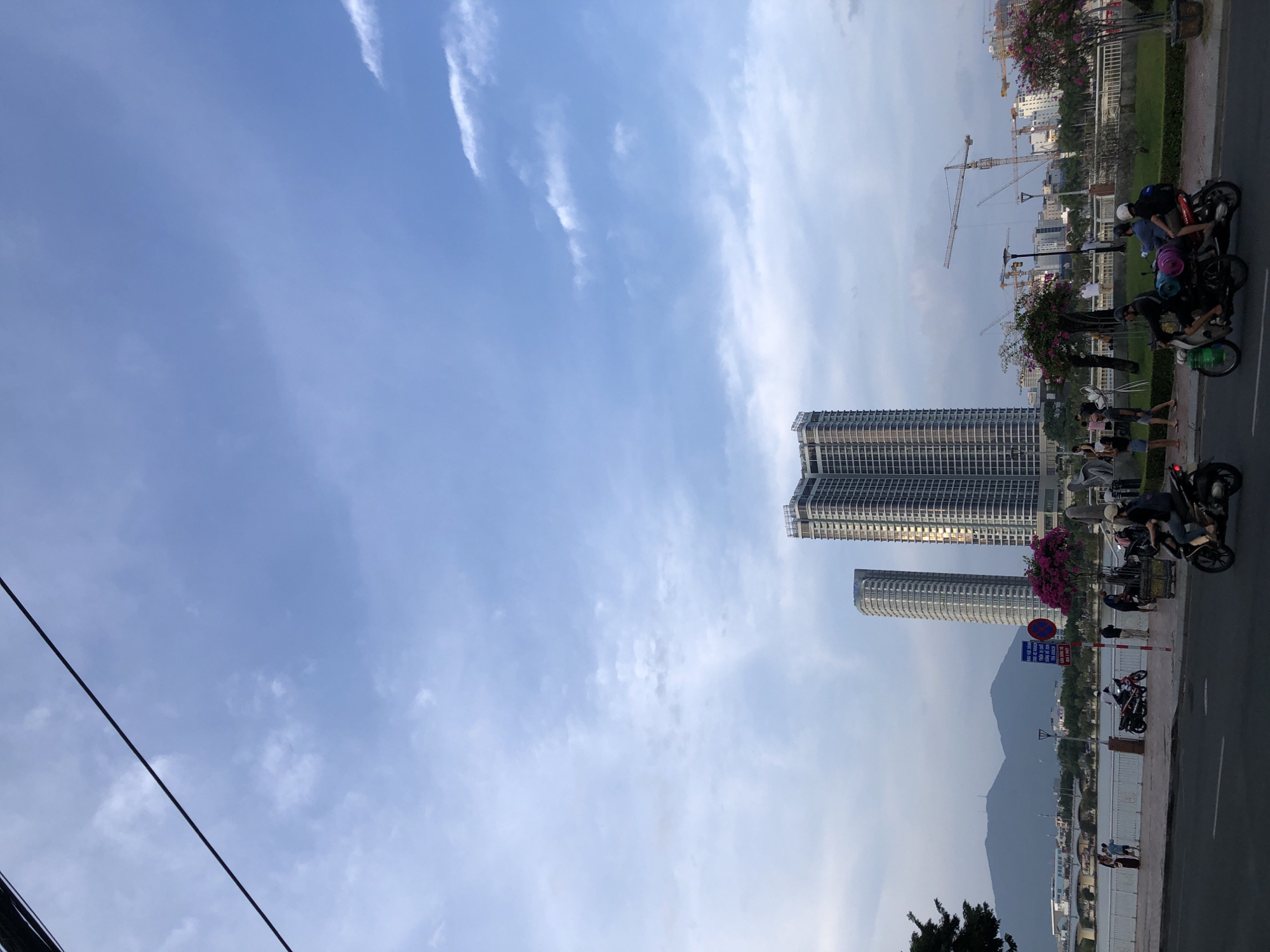 The image depicts a street scene in the city of Đà Nẵng, Vietnam, with a prominent riverfront area. Two tall, modern apartment buildings are visible in the background, with one appearing to be under construction. A river or body of water separates the foreground from the buildings and a distant mountain range. The foreground features a sidewalk lined with trees and a street where several motorbikes are in motion. People are seen walking and riding on the motorbikes. A sign with Vietnamese text is posted on a pole. The sky is cloudy, with scattered white clouds against a pale blue background. Construction cranes are also visible in the distance, suggesting ongoing development.