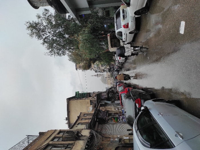 The image depicts a wet, narrow street or alleyway in what appears to be a Middle Eastern city, specifically Al-Qanawat Municipality, Syria. The scene is captured on an overcast day, indicated by the diffused light and the damp, reflective surface of the asphalt road, suggesting recent rainfall.
The street is lined on both sides with parked cars of various models and colors, including white, red, black, and silver/grey. A bicycle with a large, dark delivery bag on its rear rack is parked closer to the center of the street. Further down the street, a person wearing a light brown jacket walks away from the viewer, while another figure is visible near parked cars on the left.
On the left side of the street, older, multi-story buildings with weathered facades, some showing signs of disrepair like exposed brick and broken windows, line the street. Utility lines and cables crisscross above these structures. On the right, the buildings appear slightly more modern or better maintained. Above the rooftops in the distance, a prominent domed structure with a slender minaret, characteristic of a mosque, is clearly visible, providing a strong cultural and geographical clue. Lush green trees are interspersed among the buildings, particularly on the left.
The overall impression is one of everyday urban life in a bustling, historical area, under a quiet, rainy sky. No discernible text is visible on signs or vehicles.