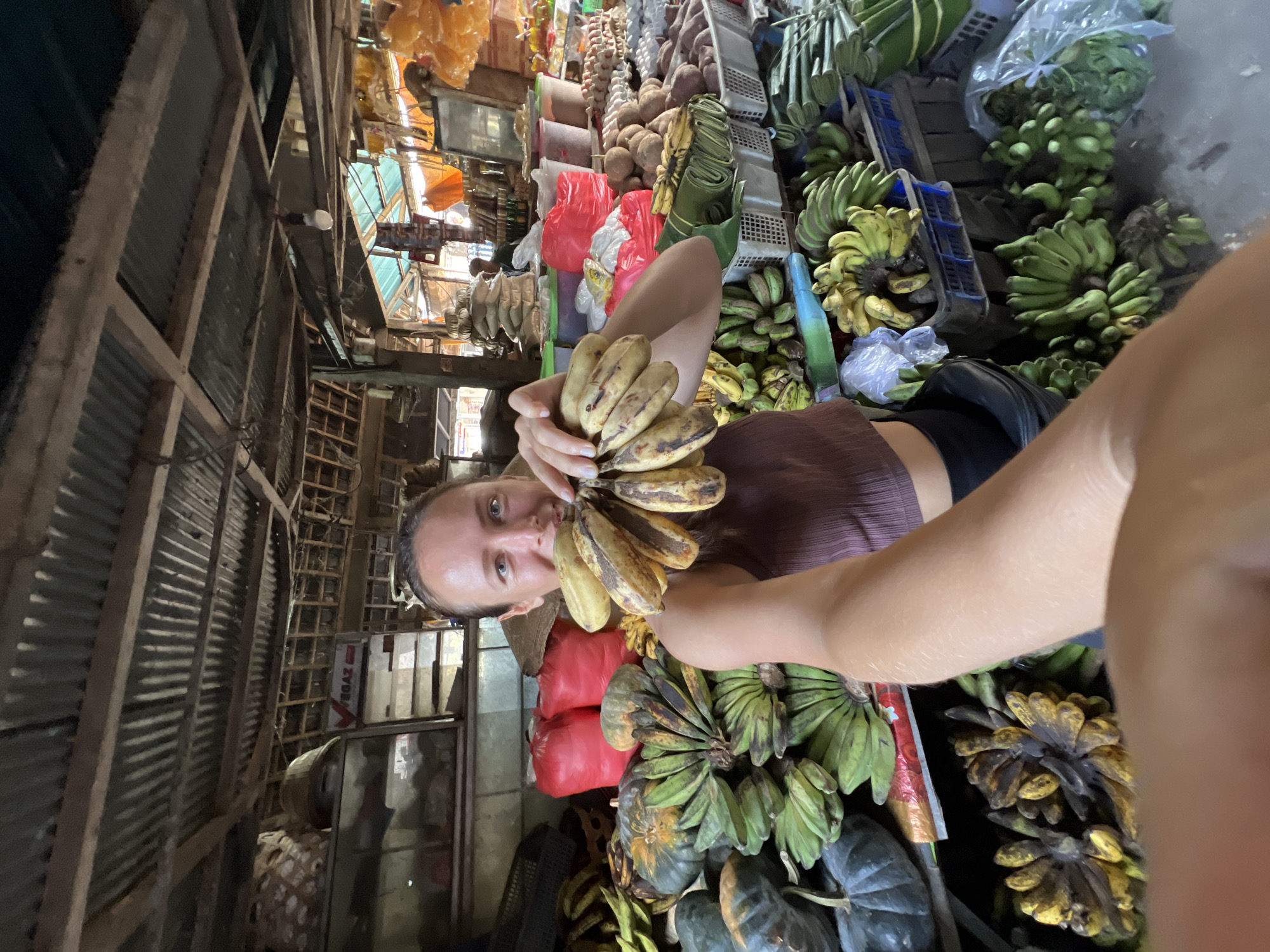 In Ubud District, Indonesia, a woman stands in a market stall, holding a bunch of bananas. She is positioned in the foreground of the image, with her arm extended and a smile on her face. The market stall is filled with various fruits and vegetables, including pumpkins and more bananas. The stall is covered with a corrugated metal roof and has wooden beams. The background is blurred but suggests a busy market environment.