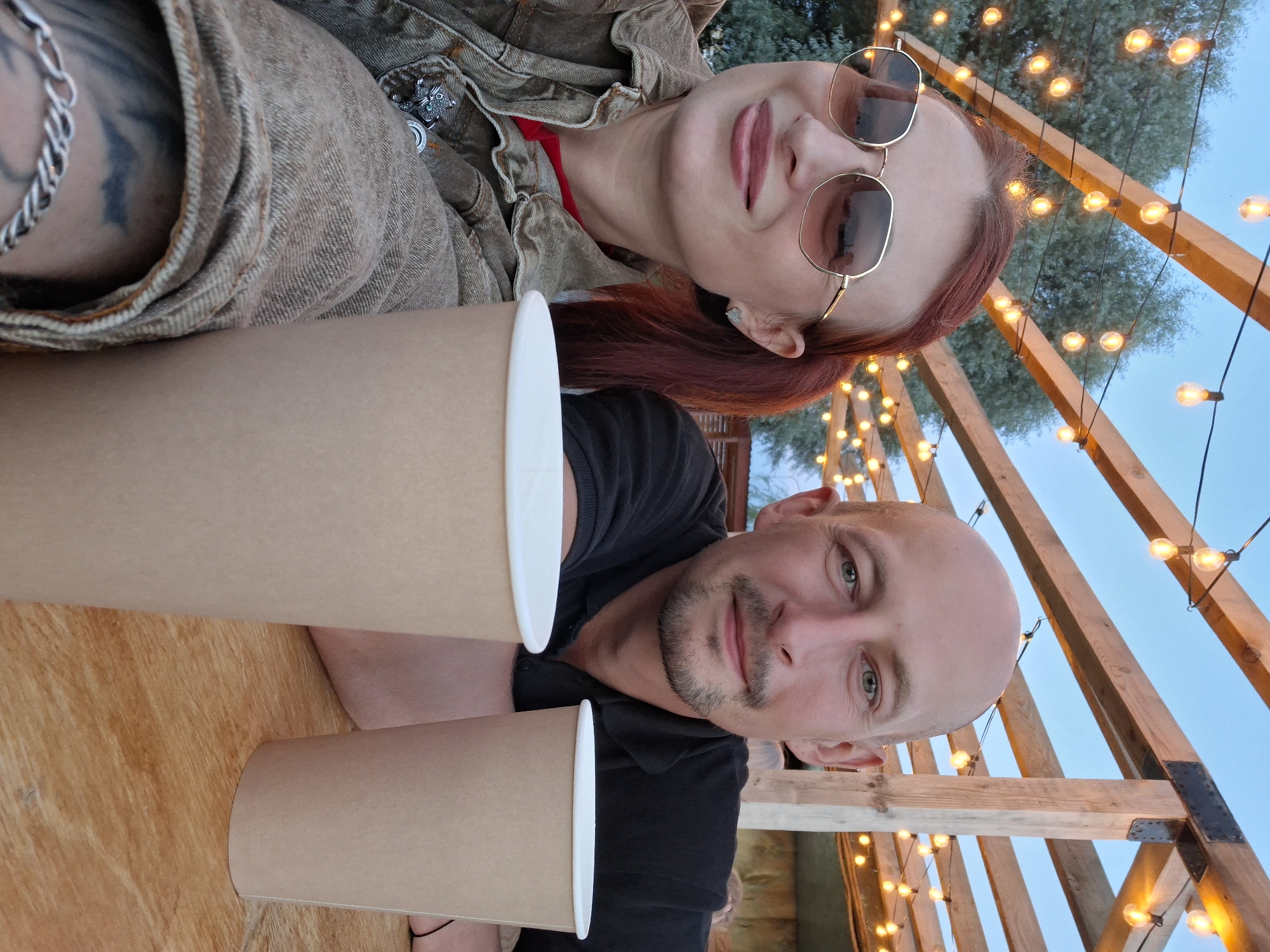 This is a selfie of a man and a woman sitting at a wooden table outdoors. The woman is on the left, wearing sunglasses and a denim jacket over a red shirt. She has red hair pulled into a ponytail. The man is on the right, wearing a black polo shirt. He is bald with a goatee and mustache. In front of them are two disposable cups. Behind them, strings of lights are strung across wooden beams, and trees are visible. The sky is a dusky blue. The location is described as being in the city of Myadzyel, Belarus.
