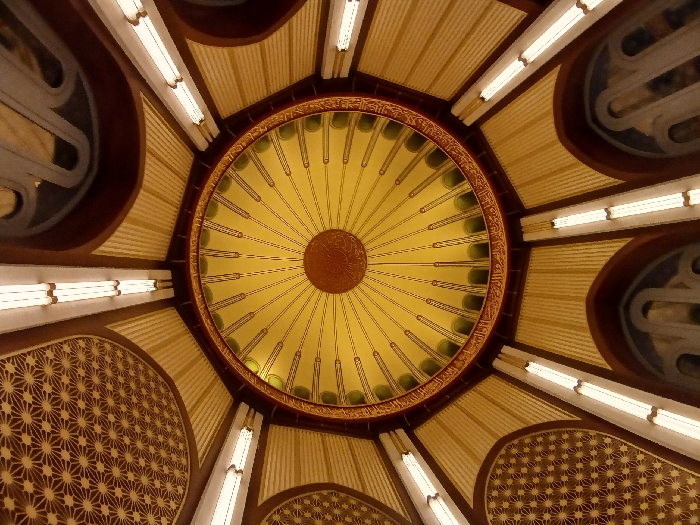 The image shows the interior of the Süleymaniye Mosque in Istanbul, Türkiye. The camera is pointed upwards towards the dome of the mosque. The dome is painted with a yellow and green pattern, with an inscription in Arabic script. The dome is illuminated by light fixtures that are mounted to the walls. The mosque's walls are painted a dark brown color and are covered in intricate geometric patterns. There is no one in the image.