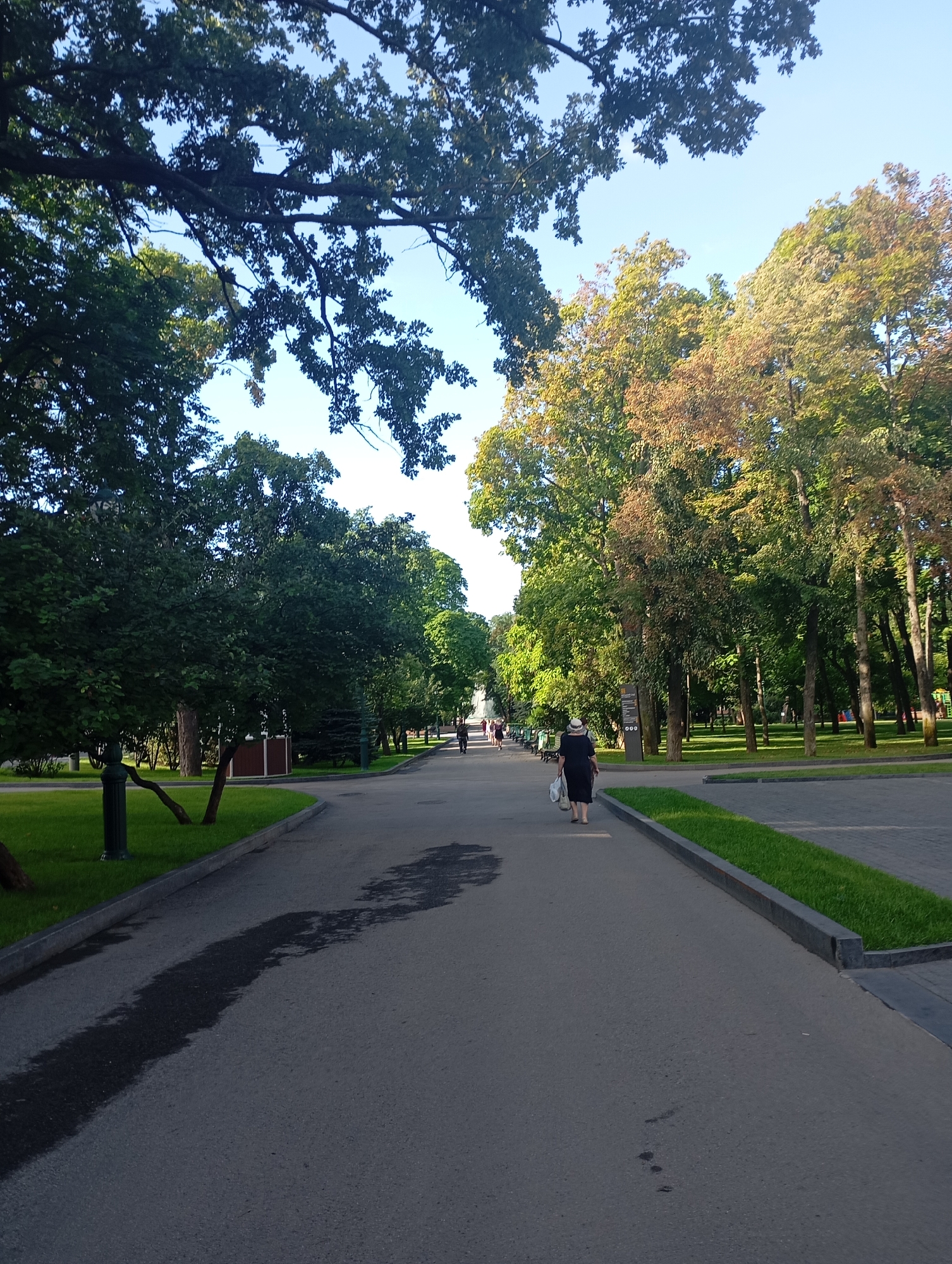 The image depicts a paved pathway in a park, lined with trees on both sides and a grassy area to the left. The pathway leads into the distance, with a few people walking along it. One person, a woman wearing a dark dress and a straw hat, is seen walking away from the camera, carrying bags. The trees vary in foliage color, with some appearing fully green and others showing hints of autumn hues. The sky is clear and blue. The setting is characteristic of a public park, likely in the city of Kharkiv, Ukraine, given the context of the request. The pathway appears to be well-maintained, with some dark, damp patches on its surface.