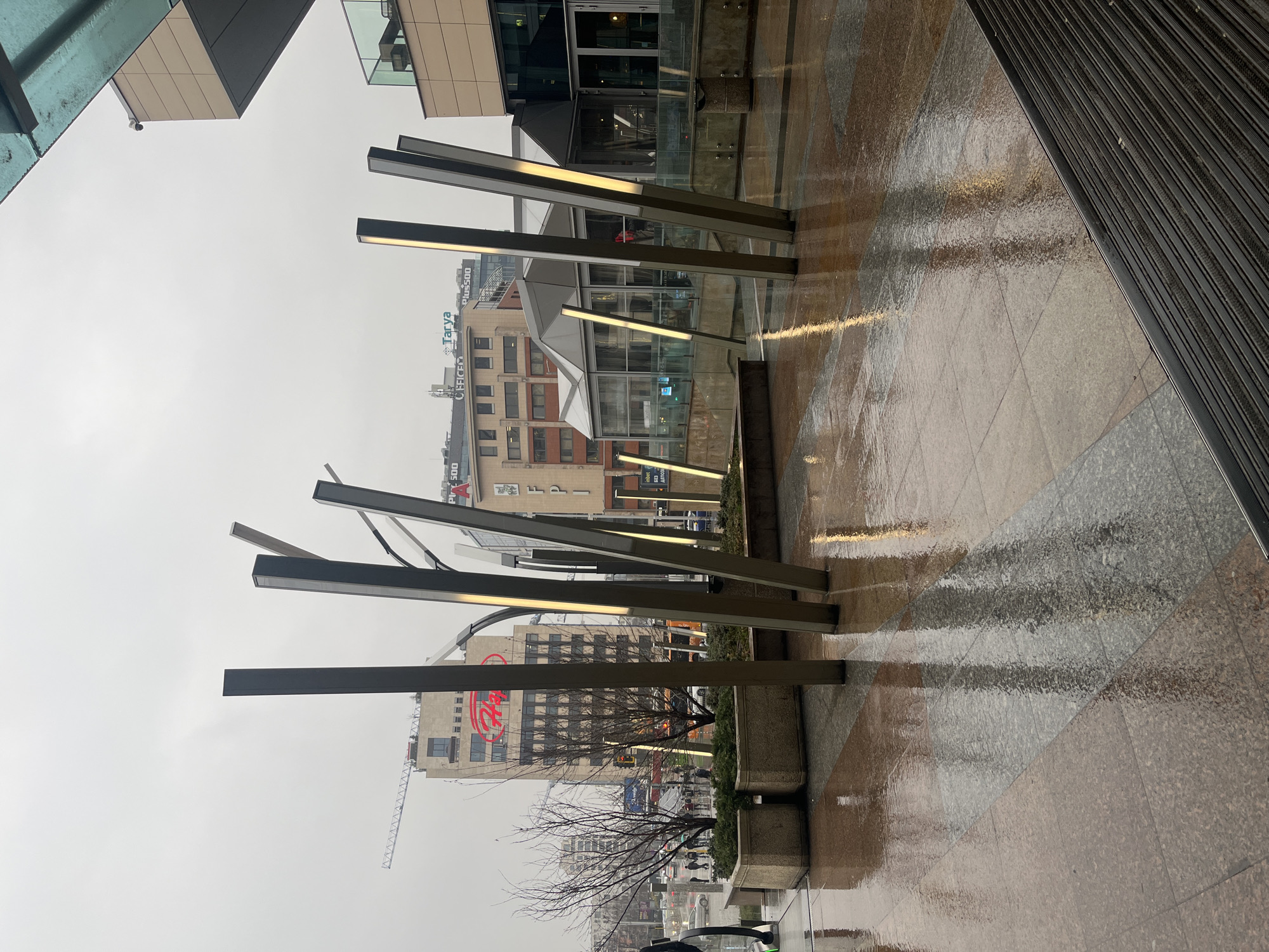 The image depicts an outdoor urban scene on a rainy day in Sofia, Bulgaria. The ground is wet and tiled, reflecting the ambient light and some of the structures above. Several vertical, rectangular metal structures with illuminated light strips are positioned along the walkway. In the background, buildings of varying heights and architectural styles are visible, including one with a prominent red "Hala" sign. A construction crane is partially visible against the overcast sky. Bare trees are present in a concrete planter along the walkway. The image is taken from a slightly low angle and is oriented in a rotated manner.