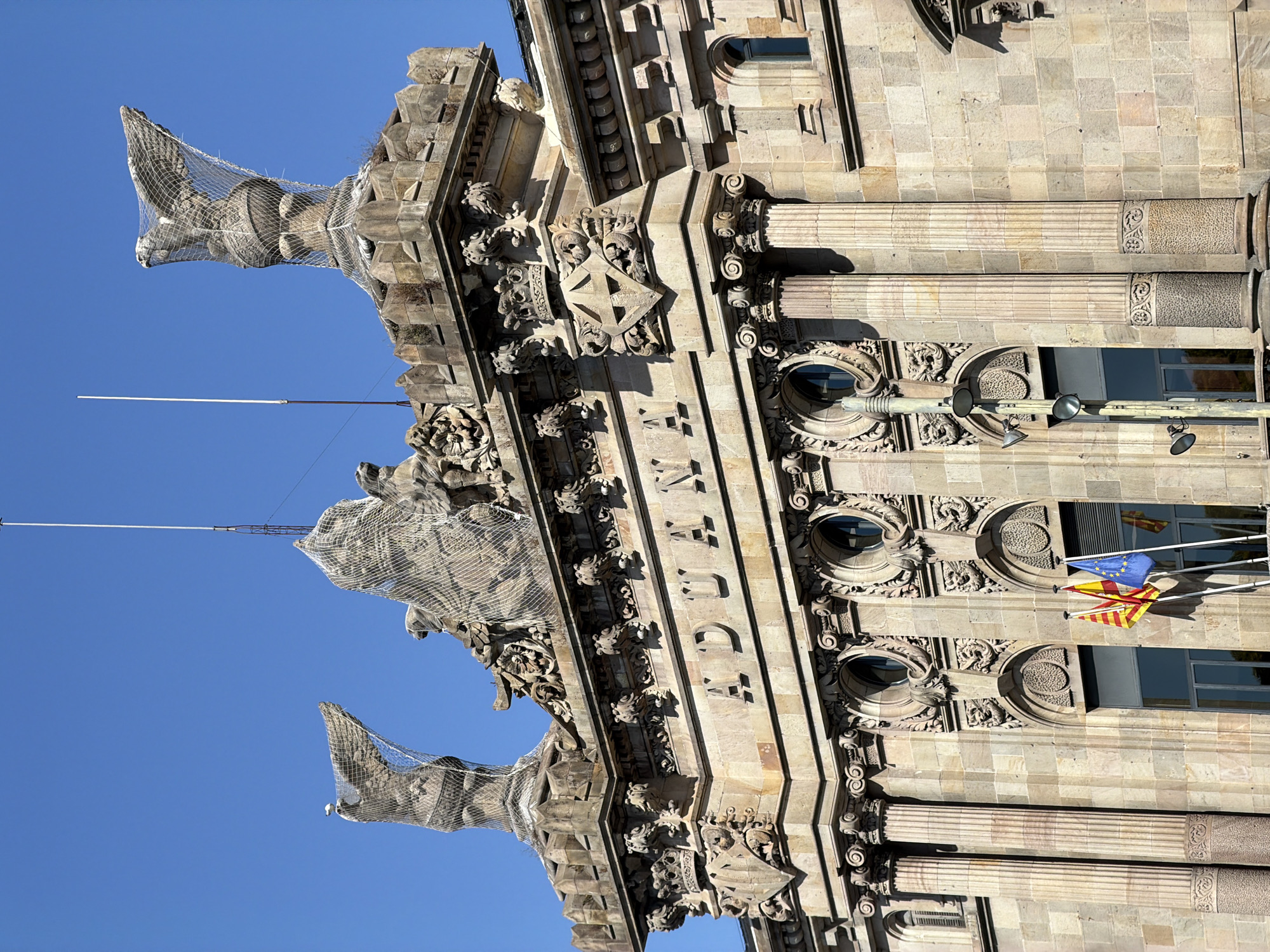 The image shows the "Aduana" building in Barcelona, Spain, under a clear blue sky. The architecture is classical, with large columns, ornate carvings, and statues adorning the rooftop. Specifically, two large eagle statues, covered by protective netting, are visible. Below them, the word "ADUANA" is inscribed in large stone letters across the building's facade. Three flags, the Catalan flag, the Spanish flag, and the European Union flag, are prominently displayed at the lower portion of the building, indicating their presence in Spain. The building's facade is constructed from light-colored stone, showing intricate details in the stonework. The environment is bright and sunny, casting distinct shadows.