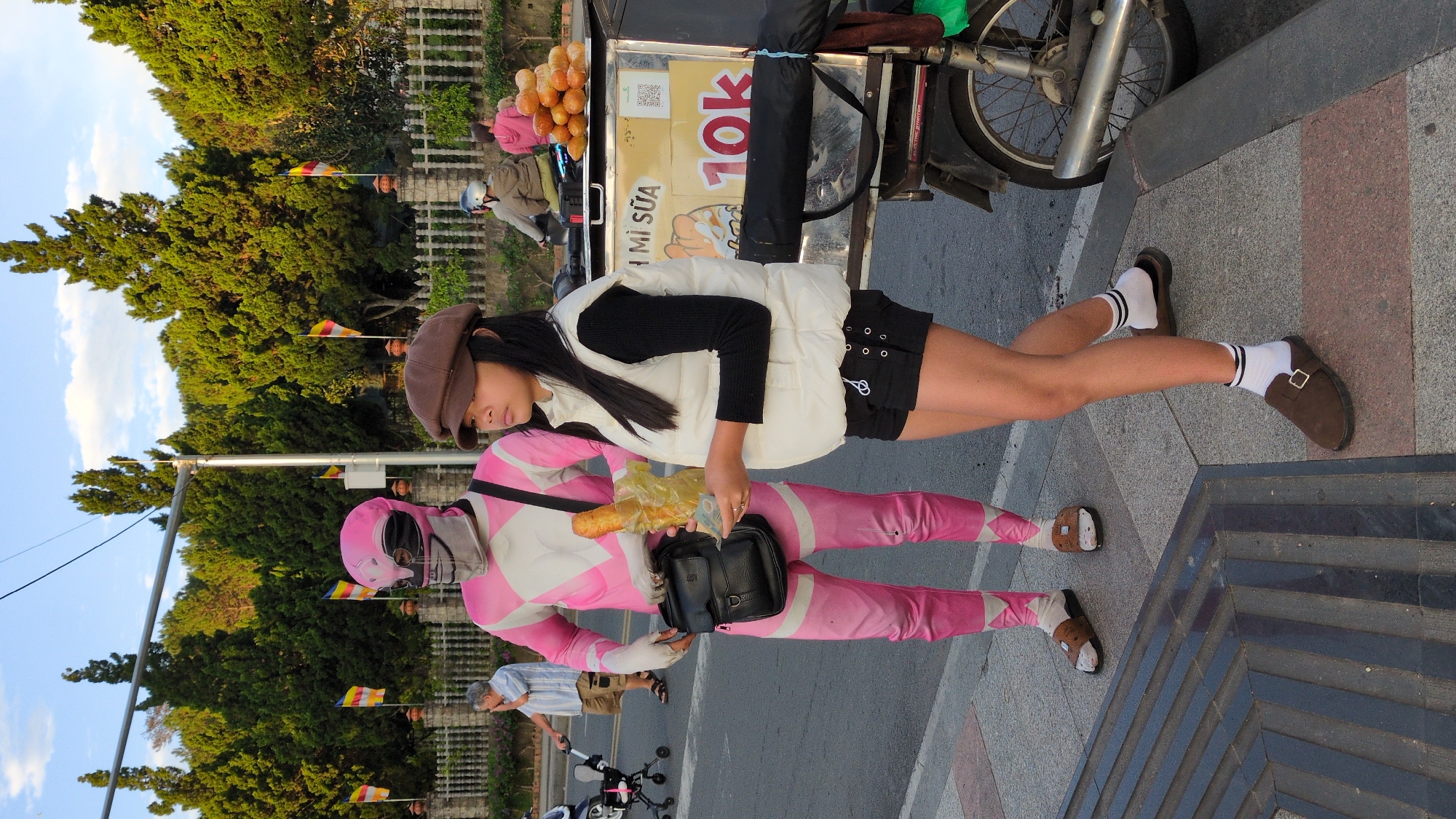 In the city of Da Lat, Vietnam, a person in a pink Power Ranger costume stands next to a young woman on a sidewalk. The Power Ranger is holding a baguette wrapped in plastic and a small stack of currency. The woman, wearing a white vest over a black long-sleeved shirt and black shorts, is also holding a baguette in a similar plastic wrap. They are positioned in front of a food cart selling various pastries. The food cart has a sign with Vietnamese text and the number "10k" prominently displayed, likely indicating a price. Behind them, a metal fence separates the sidewalk from a street lined with trees. Several flags, some colored yellow, red, and white, are visible on poles near the fence. In the background, a man in a light-colored shirt pushes a stroller, and other individuals are seen on a motorcycle, some wearing helmets. The overall scene depicts a street-level view with everyday urban activity and a touch of playful costume.