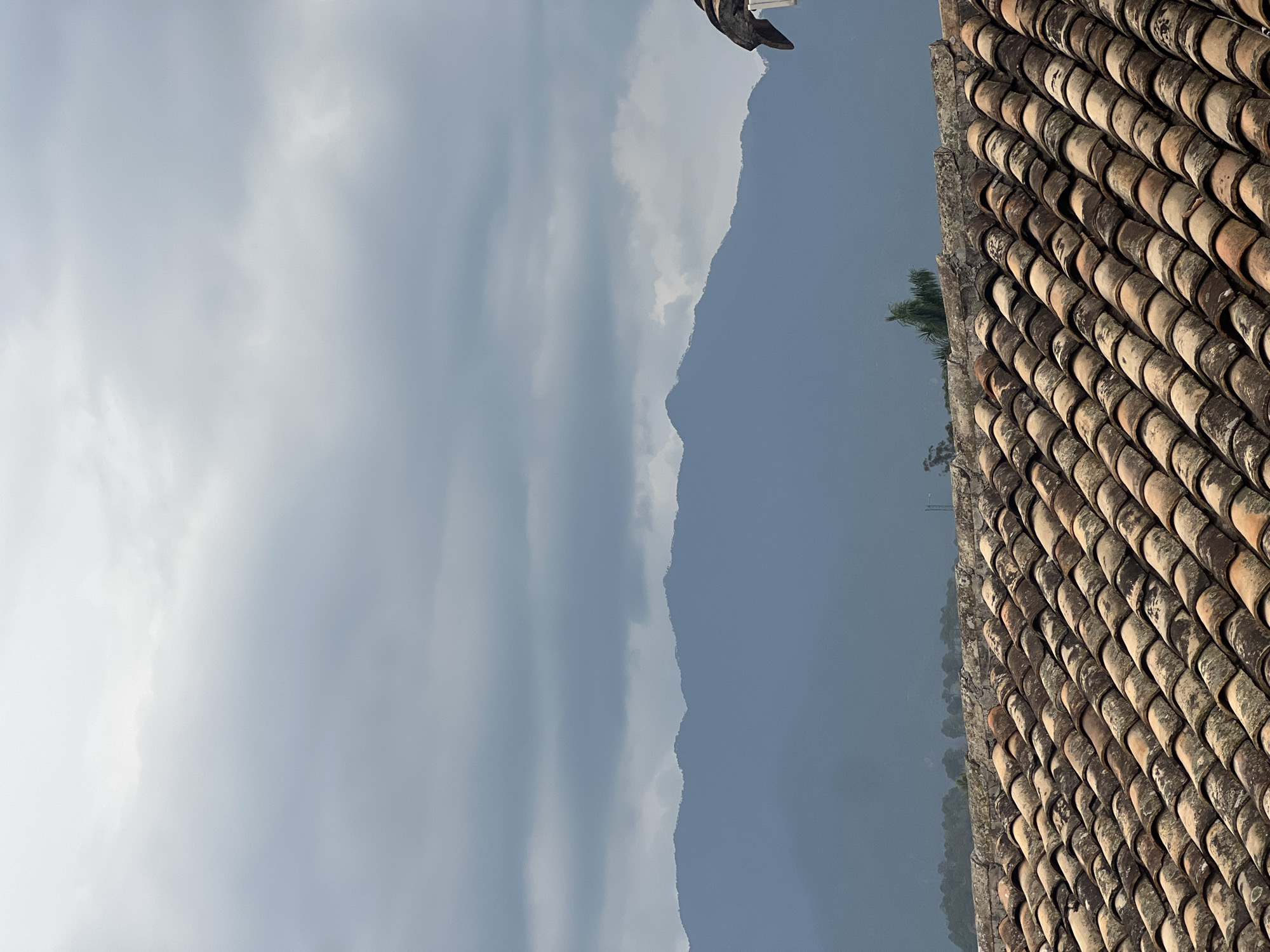 A partially obscured view from a rooftop in Gracias, Honduras, with the tiled roof of a building taking up the right side of the frame. The foreground tiles are a weathered orange-brown color, running in parallel lines. Beyond the roof, the middle ground features a muted blue mountain range under a cloudy sky. The left side of the image shows a broad expanse of pale gray clouds, with the sky and mountain range partially obscured by haze.