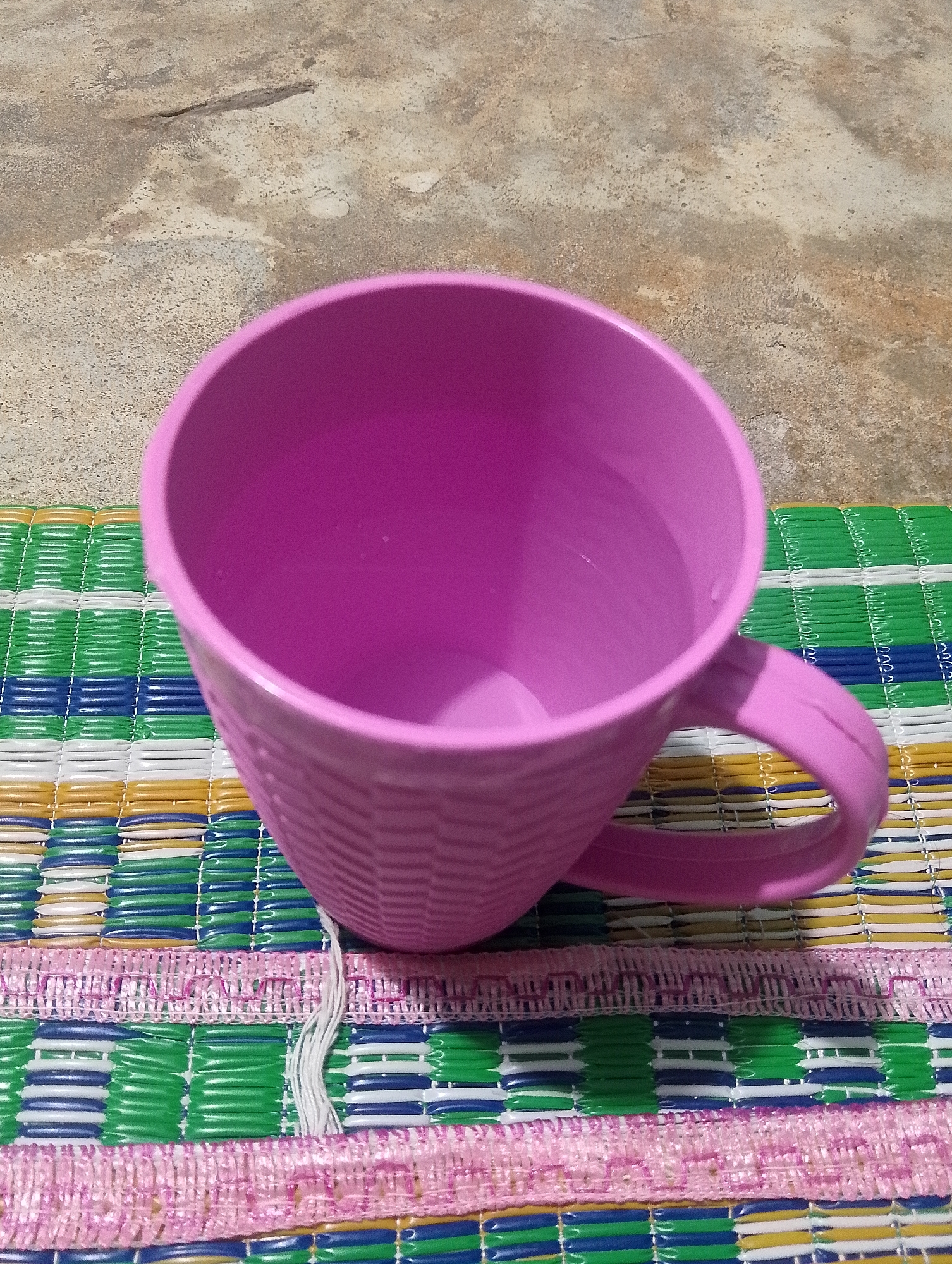 A pink plastic mug filled with clear liquid rests on a woven mat. The mug has a textured exterior, resembling a basket weave pattern. The mat is colorful, with alternating stripes of green, white, yellow, and blue. Two pink fabric borders with white threads are visible at the bottom of the mat. The ground behind the mat appears to be a rough, light-colored concrete surface. The scene is outdoors, under natural light.
