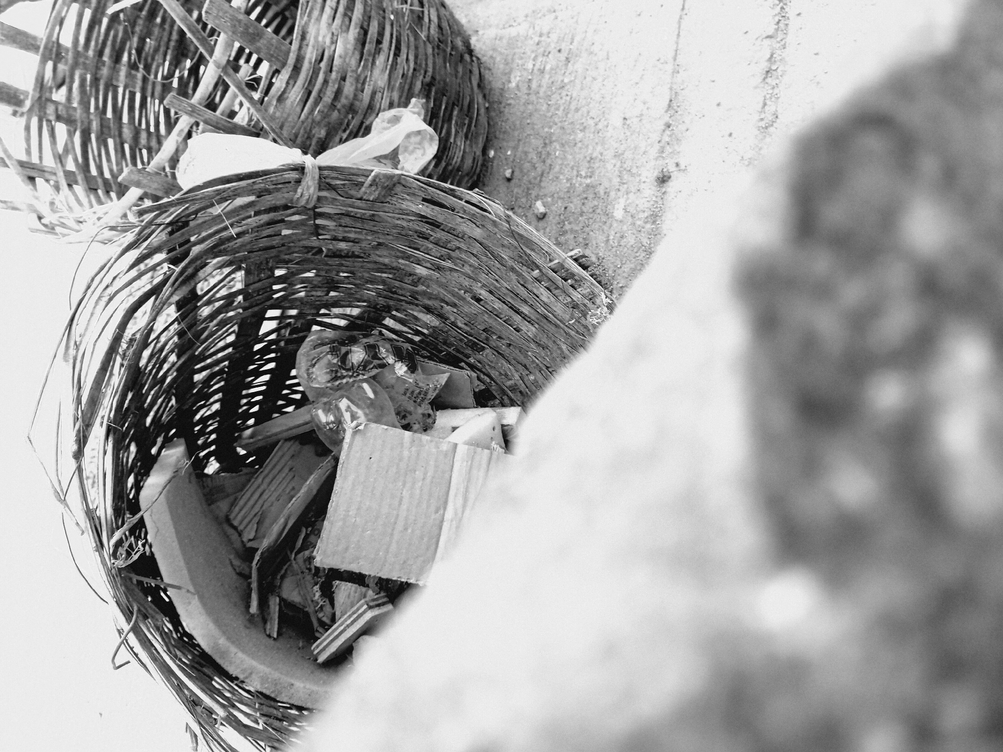 The image shows two wicker baskets filled with trash, likely situated on a concrete surface. The lower basket is full of cardboard and plastic debris. The second basket, stacked on top, also holds trash. The blurry right edge of the frame shows what seems like a piece of a large, rough-textured structure. While the location is specified as Bukuru, Nigeria, there are no specific localizing factors such as landmarks, clothing, or visible signage to confirm this. The scene depicts discarded refuse and lacks signs of human presence or activity.