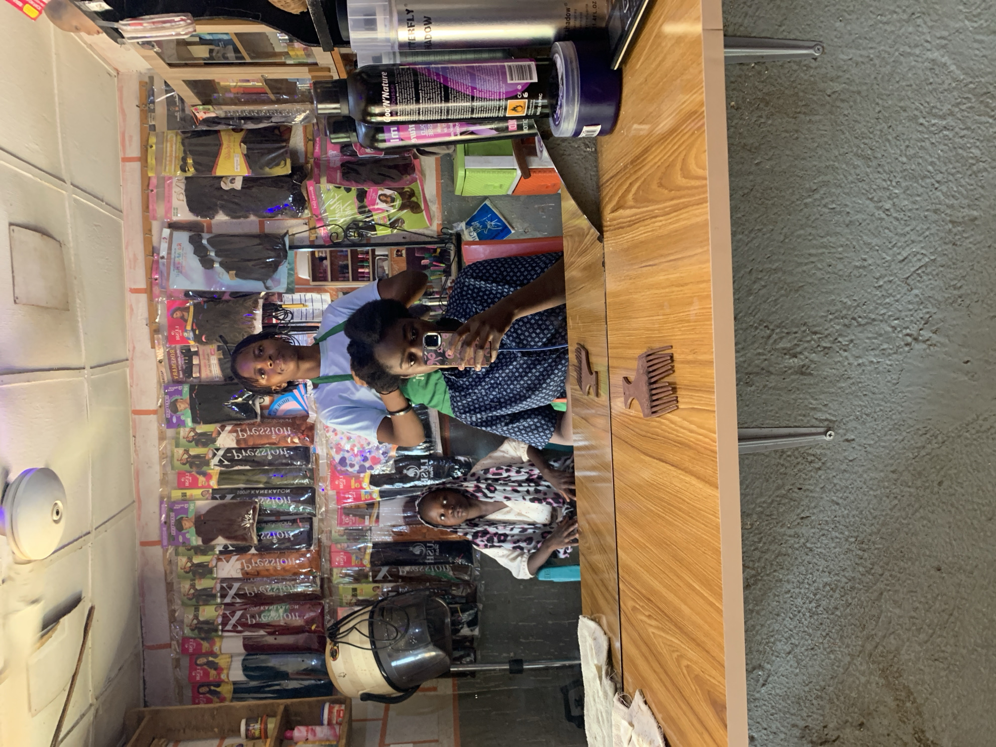 The image depicts a scene inside what appears to be a hair salon in Rafin Par, Nigeria. Two women are visible; one is likely a stylist, seen partially, wearing a light blue shirt and a green wristband. She is styling the hair of another woman who is holding a cell phone, possibly taking a photo or video. This woman is wearing a patterned blue and white shirt. Another woman wearing a white patterned scarf is sitting in a chair looking straight into the mirror.
The environment shows hair extensions are displayed on the wall. Above the stylist and customer are various hair products on a shelf, including containers labeled "Good 'N Nature" and "BUTTERFLY Shadow". The mirror reflects the scene, and a comb is visible on the surface of the table. A hair dryer is near the bottom of the reflection. The ceiling and wall are visible on the left side of the photo. The wall on the right is grey and there are two metal rods attached.