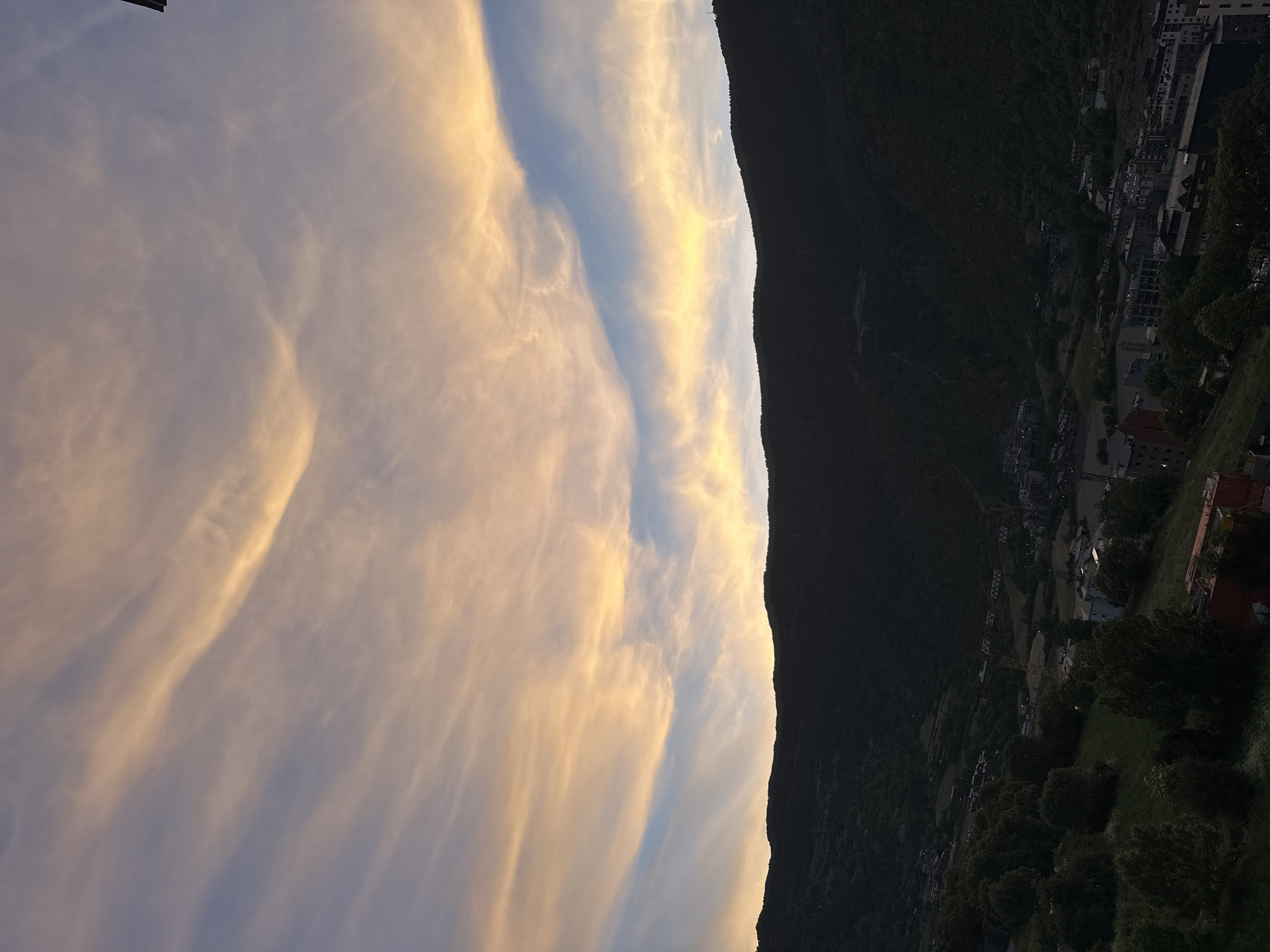 This image depicts a serene mountain landscape at **twilight**, most likely **sunset**, over a valley town identified as Encamp, Andorra. The sky, which occupies the majority of the frame, is a canvas of soft, ethereal clouds. Long, wispy cirrus-like clouds are illuminated with warm hues of golden yellow, soft orange, and delicate pink, reflecting the sun's low angle. A distinct bright band of golden light stretches horizontally across the middle of the sky, indicating where the last direct light of day touches the clouds, gradually fading to a paler blue in other areas.
To the right, a dramatic, dark **mountain range** rises, creating a strong silhouette against the luminous sky. Its rugged peaks and slopes are shrouded in deep shadow, hinting at dense tree cover. Nestled within the valley at the base of these mountains, and extending into the lower right of the image, is a **town**. The buildings are largely in shadow, appearing dark, though some faint lights are visible, suggesting the encroaching darkness. Patches of dark green trees and vegetation are scattered throughout the valley and on the lower mountain slopes. A **construction crane** can be faintly seen among the town's structures in the mid-ground. The scene evokes a sense of tranquil beauty as daylight fades, painting the sky with rich colors over the quiet mountain settlement. No visible text is present.