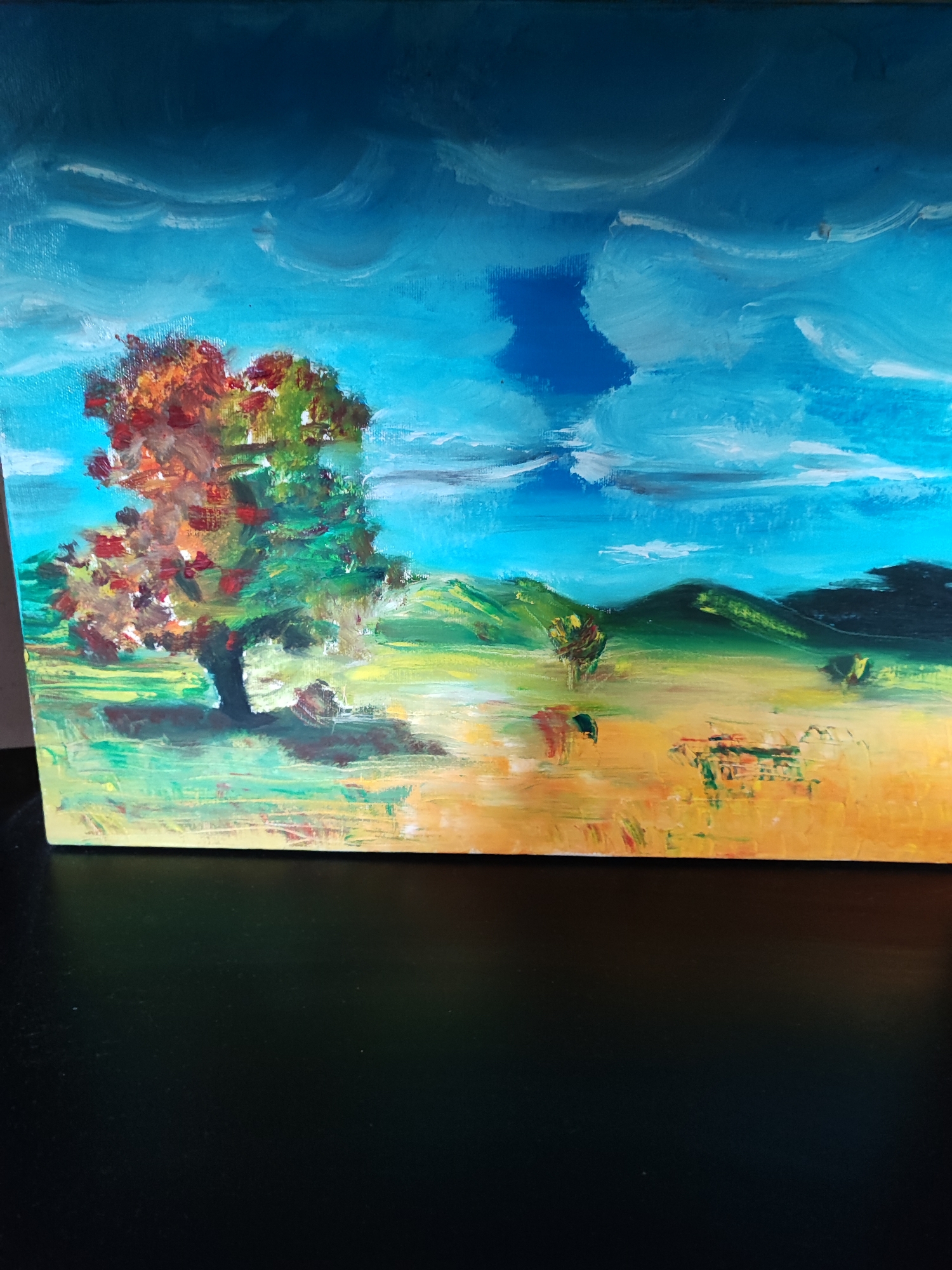 A vibrant acrylic landscape painting depicts a pastoral scene. The depicted location is stated to be within the city of Kadievo, Bulgaria. The composition features a bright blue sky with dynamic white and light blue clouds, darkening towards the top. A prominent tree with a thick, dark trunk and foliage in a mix of autumnal reds, oranges, and greens stands in the left foreground, casting a shadow to its right. The foreground transitions into a broad field rendered in warm yellows and greens, leading to rolling hills in various shades of green, with darker, more distant hills against the horizon. Smaller, less defined trees are scattered across the middle ground field. The brushwork is visible and expressive, creating texture and movement throughout the depiction of open terrain under a vivid sky.