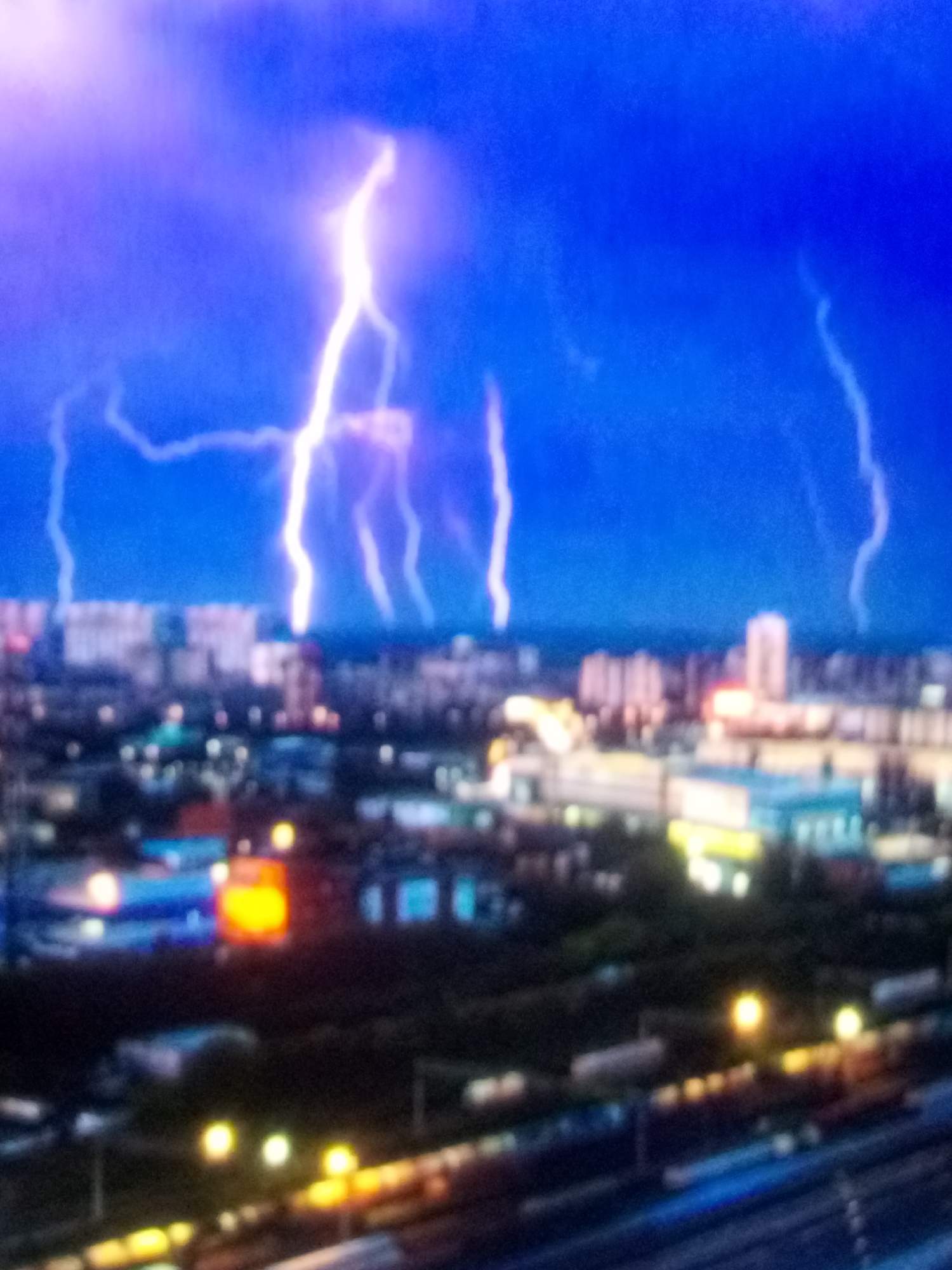 A wide-angle, blurred, aerial shot captures a city skyline at night under a dark blue sky illuminated by multiple bright white lightning strikes. The lightning bolts are jagged and branch out as they descend towards the cityscape. The city below is a blur of lights, with illuminated buildings of varying heights and some distinct colored lights, including orange and blue. In the foreground, train tracks and the tops of what appear to be train cars or freight can be seen, also dotted with lights. The overall scene depicts a powerful storm over an urban environment.
