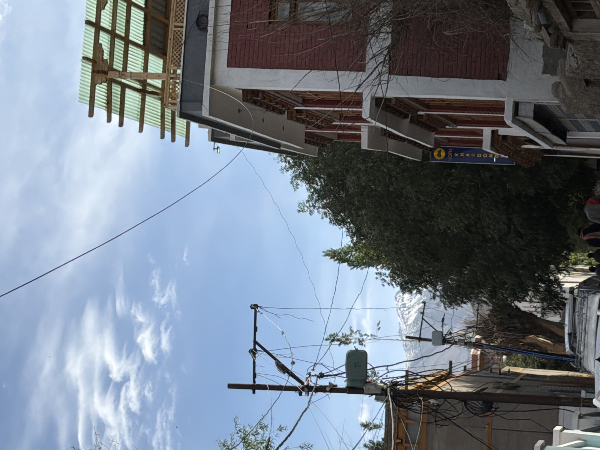 The image depicts a street scene in Leh, India. The primary focus is a multi-story building with red brick and white accents, featuring traditional architectural elements like carved wooden eaves. A blue sign attached to the building reads "HOTEL KHARDONG". Above the building, a corrugated green roof is supported by wooden beams. On the left side of the image, a utility pole is heavily laden with a dense network of electrical wires and cables, with a green junction box attached. Trees are visible, including a large, dark green tree behind the hotel and a bare tree trunk to the left of the pole. In the background, snow-capped mountains rise against a blue sky with scattered white clouds. A few people are visible walking in the street, and the roof of a white vehicle is seen in the foreground. The overall impression is of a bustling urban environment nestled within a mountainous landscape.