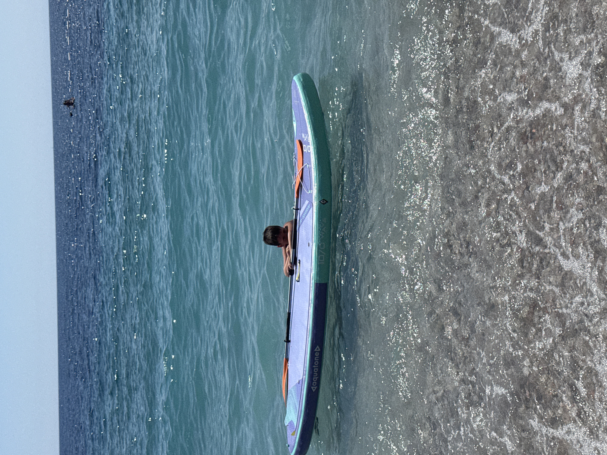 A young boy, his head and shoulders visible, rests on a stand-up paddleboard in the shallow, clear waters off the coast of Mirište, Montenegro. The paddleboard, a vibrant blue and teal with "aquatone" branding, floats on the surface. An orange paddle rests on the board. The boy is looking down towards the paddleboard. In the distance, the vast expanse of the Adriatic Sea stretches out, meeting a clear blue sky. The sun glints off the water's surface, creating a shimmering effect. The seabed is visible through the clear water, showing pebbles and sand.