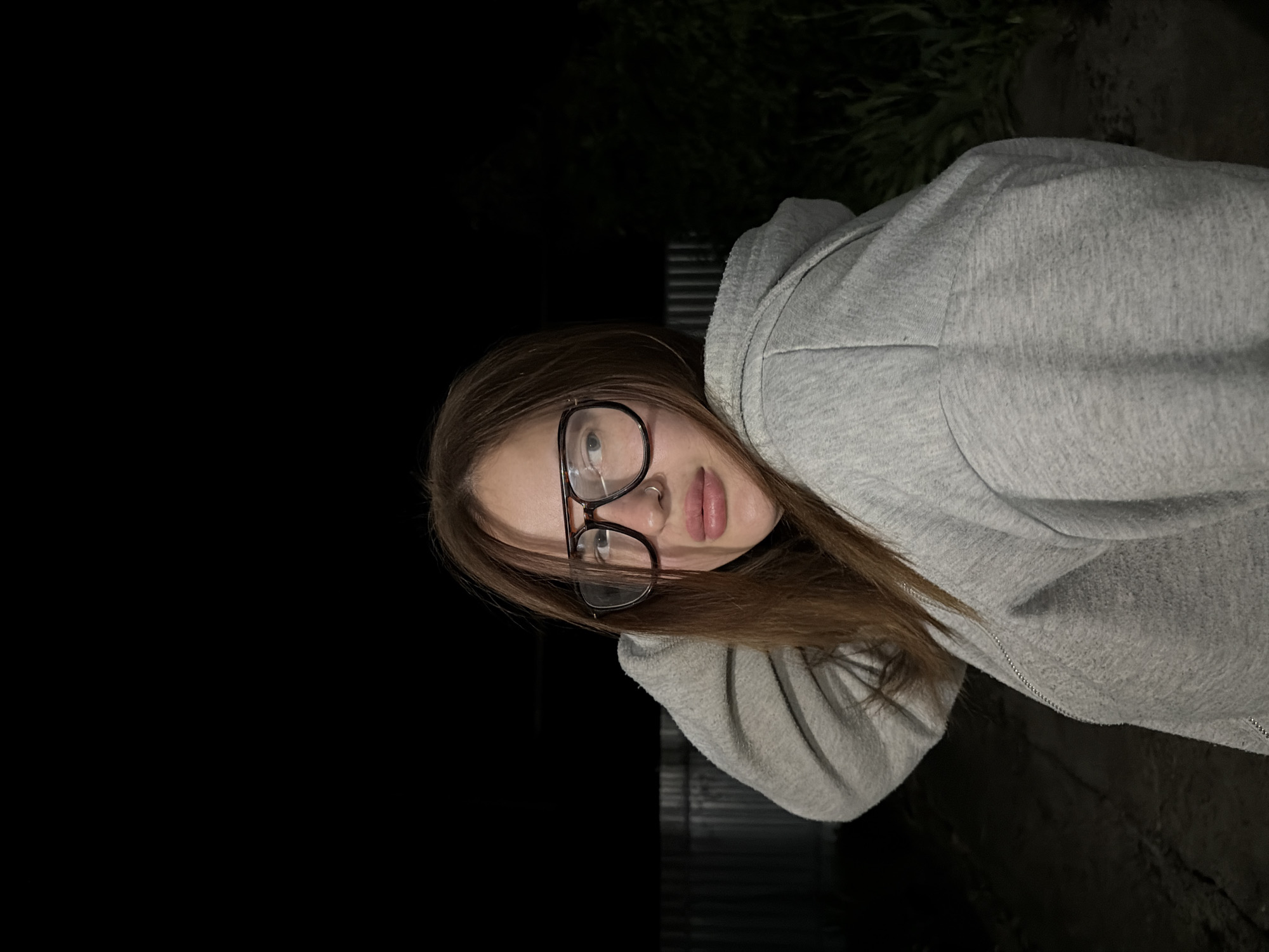 The image depicts a young woman with light brown hair and fair skin, wearing eyeglasses with dark frames and a silver nose ring. She is wearing a gray hoodie. The background is primarily dark, suggesting it is nighttime. Some greenery is visible in the upper right portion of the image, with a fence in the background. The location is Novopokrovskaya, Russia. There are no visible actions or interactions taking place in the image.