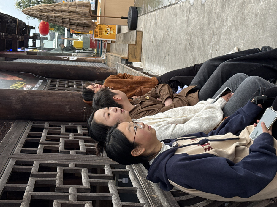 Four individuals are seated on a wooden bench against a traditional dark wooden building facade in Pengshui Miao and Tujia Autonomous County, China. From left to right: a female wearing glasses and a navy and beige hooded sweatshirt, holding a smartphone in a clear case; a female in a light-colored knitted sweater; a male in a brown collared shirt with arms crossed; and a female wearing glasses and a brown fuzzy jacket, leaning back. All individuals appear to be looking to their right, off-frame. The building features dark wooden lattice panels and vertical wooden pillars. The ground in front of the bench is a wet, grey paved surface. In the background, a red Chinese lantern is visible, along with a conical thatched parasol on a black base, and a yellow sign featuring a cartoon tiger. The overall environment suggests an urban or town setting, with overcast weather.
