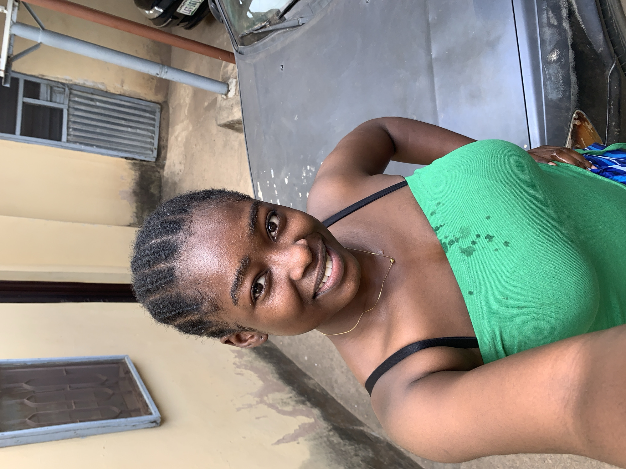 A young woman with braided hair smiles at the camera, wearing a green top with black straps and a gold necklace. She stands beside the hood of a dark-colored car. Behind the car, a beige building features a small window with dark bars. A silver metal post is visible near the building. The location is Rafin Par, Nigeria.