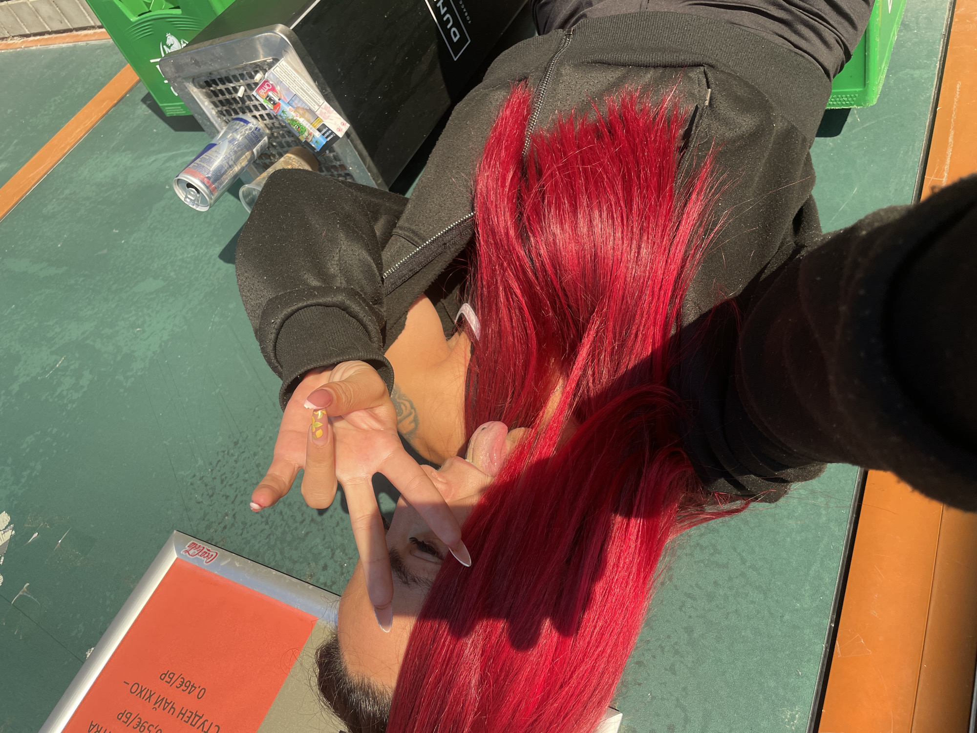 A person with bright red hair is lying down on a green surface, possibly a table or outdoor seating area. They are wearing a black zip-up hoodie. Their left hand is raised, fingers splayed in a peace sign, with a manicure featuring yellow flowers on the nails. Their right hand is partially visible, reaching towards their face. The person's face is visible, with their tongue sticking out slightly.
In the background, there is a green recycling bin and a black object with the word "DUN" visible, possibly part of a trash can or cigarette disposal unit. A crushed aluminum can, a cigarette butt, and some papers are in a metal tray attached to the bin. A red and white Coca-Cola branded sign is also visible.
The scene appears to be outdoors during the daytime, indicated by the bright sunlight and shadows cast on the surface. The overall mood seems playful and casual. The text visible on the Coca-Cola sign and other papers suggests a commercial setting, perhaps a cafe or public space. The Cyrillic text on the sign also aligns with the location context of Sofia, Bulgaria.