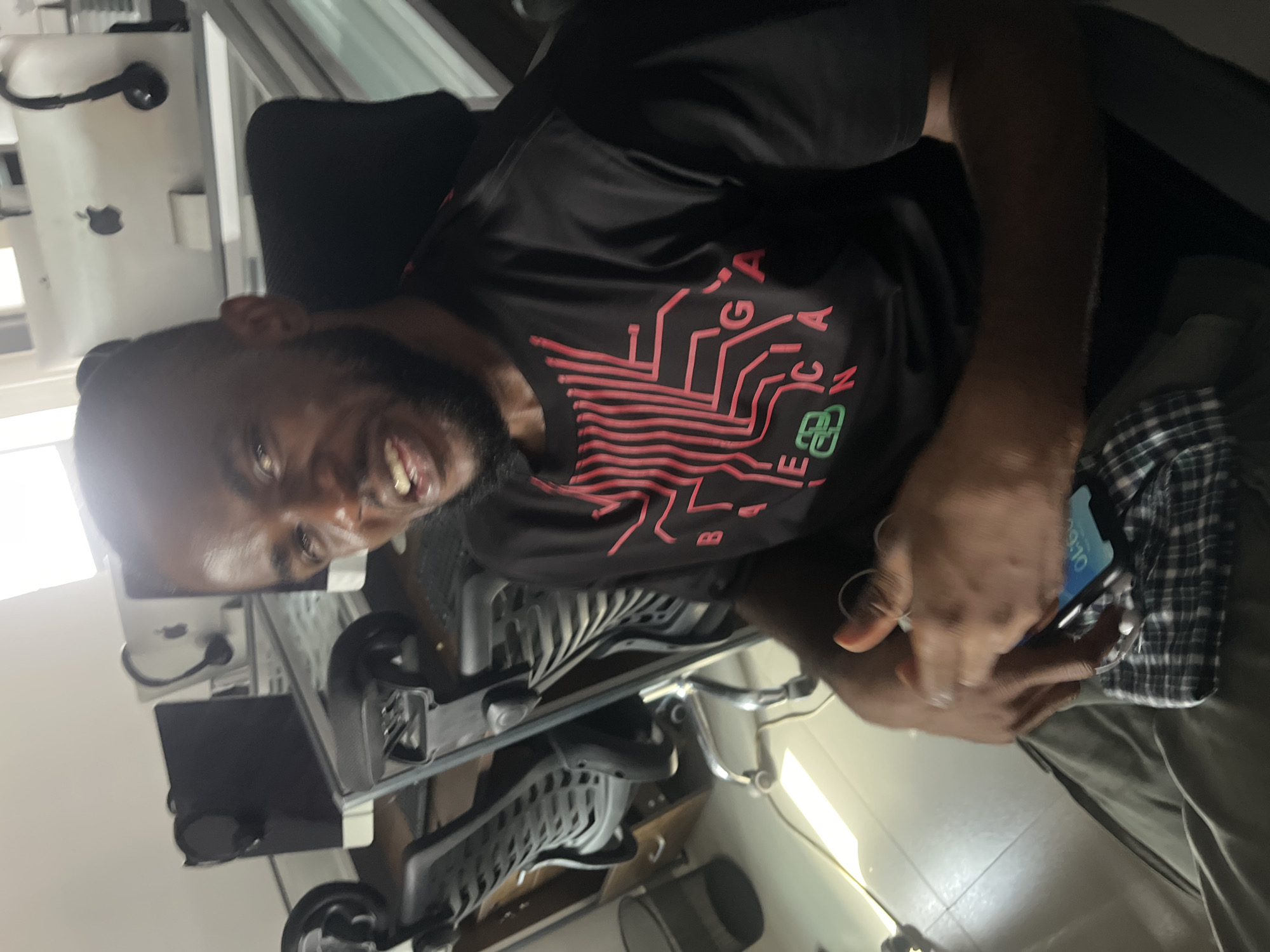 A man is sitting in a chair. He has a beard and is wearing a black t-shirt with a circuit board design on the front. He is holding a cell phone in his hands. The background includes a desk with a computer and other items. The image was possibly taken in an office in Karum, Nigeria.