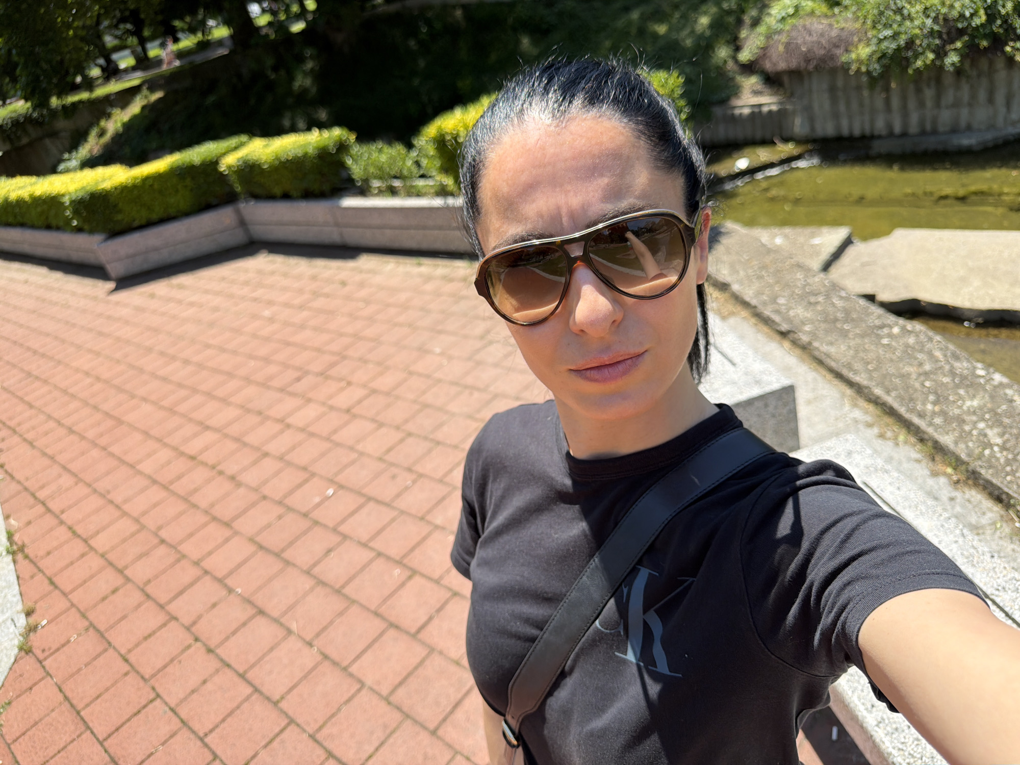 A woman is taking a selfie in a park-like setting. She has dark hair pulled back, wears large aviator sunglasses, and a black t-shirt with a grey logo. A black cross-body bag strap is visible. She stands on a brick-paved area. Behind her is a low hedge, a water feature with a concrete edge, and various trees. The exact location is difficult to pinpoint within Pleven, Bulgaria without further identifying features.