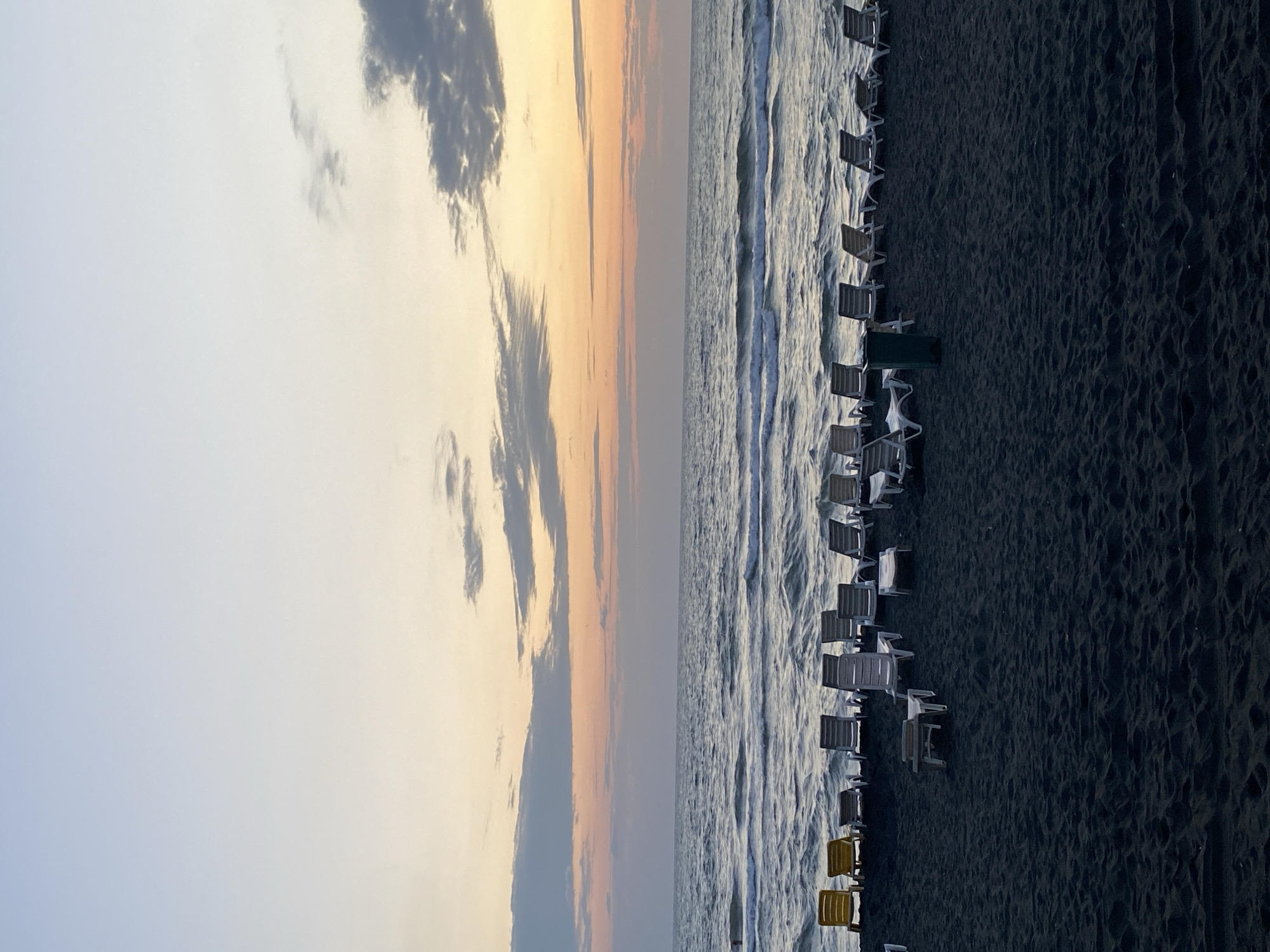 The image depicts a tranquil beach scene at Ureki, Georgia, with the Black Sea stretching out under a gradient sky of pale blue and soft pink hues, indicative of either sunrise or sunset. The foreground is dominated by dark, fine sand, characteristic of Ureki's magnetic sand beaches, with a sparse scattering of empty white and gray lounge chairs arranged in rows facing the sea. Two yellow lounge chairs are positioned further to the left. A green trash receptacle stands near the center of the beach, between two rows of lounge chairs. Gentle waves break on the shore, and a few faint ripples are visible on the water's surface. The overall atmosphere is serene and uncrowded, suggesting a quiet moment by the sea.