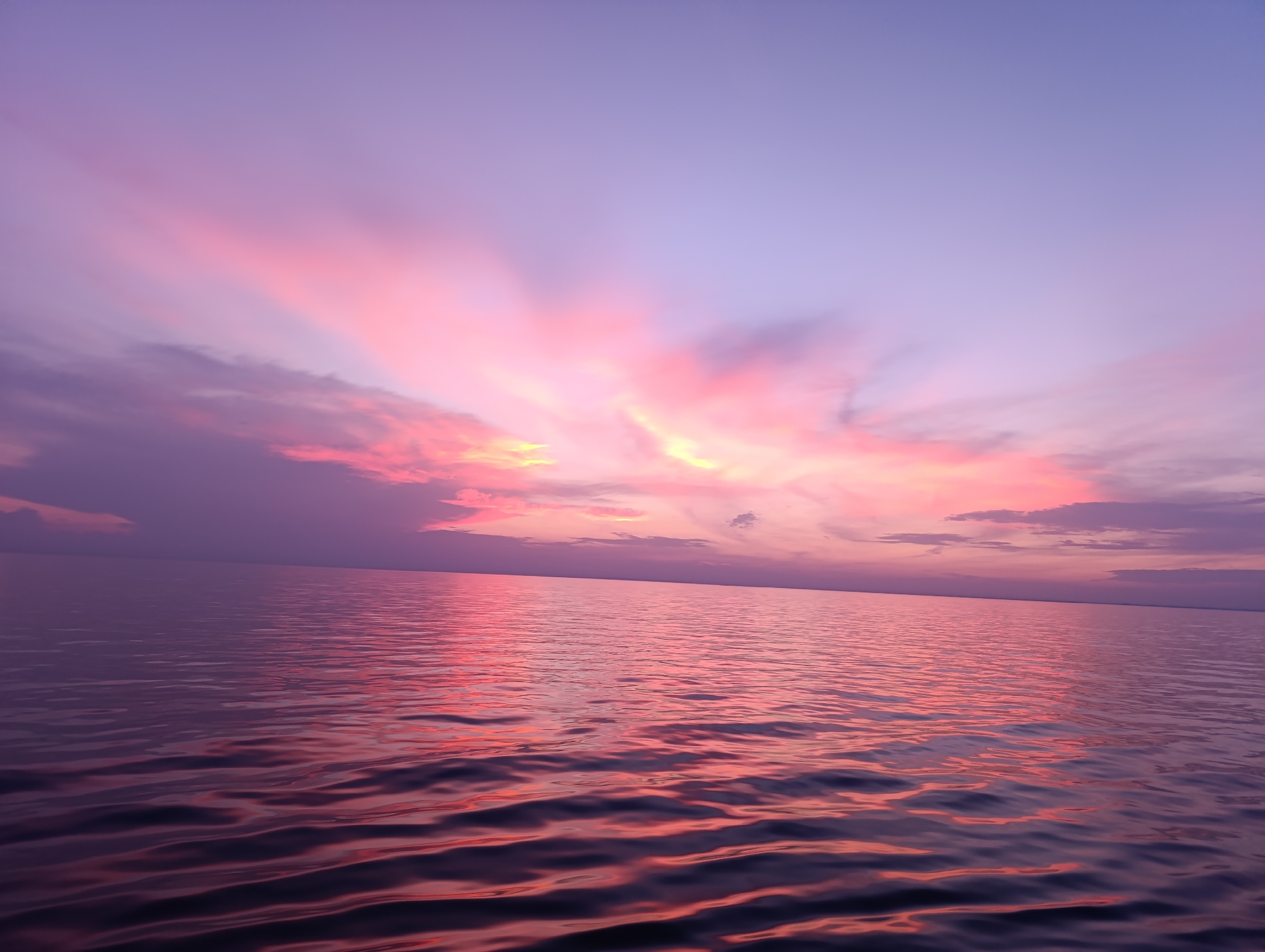The image depicts a serene seascape with a sky filled with soft hues of pink, purple, and yellow, indicative of a sunset or sunrise. The clouds are dispersed, with some appearing wispy and others more defined, illuminated by the fading light. The sunbeams fan out from behind the clouds, creating a dramatic effect. The calm water reflects these colors, with gentle ripples distorting the reflections. The horizon line is clearly visible, separating the sky from the vast expanse of water. The overall atmosphere is peaceful and tranquil, evoking a sense of natural beauty.