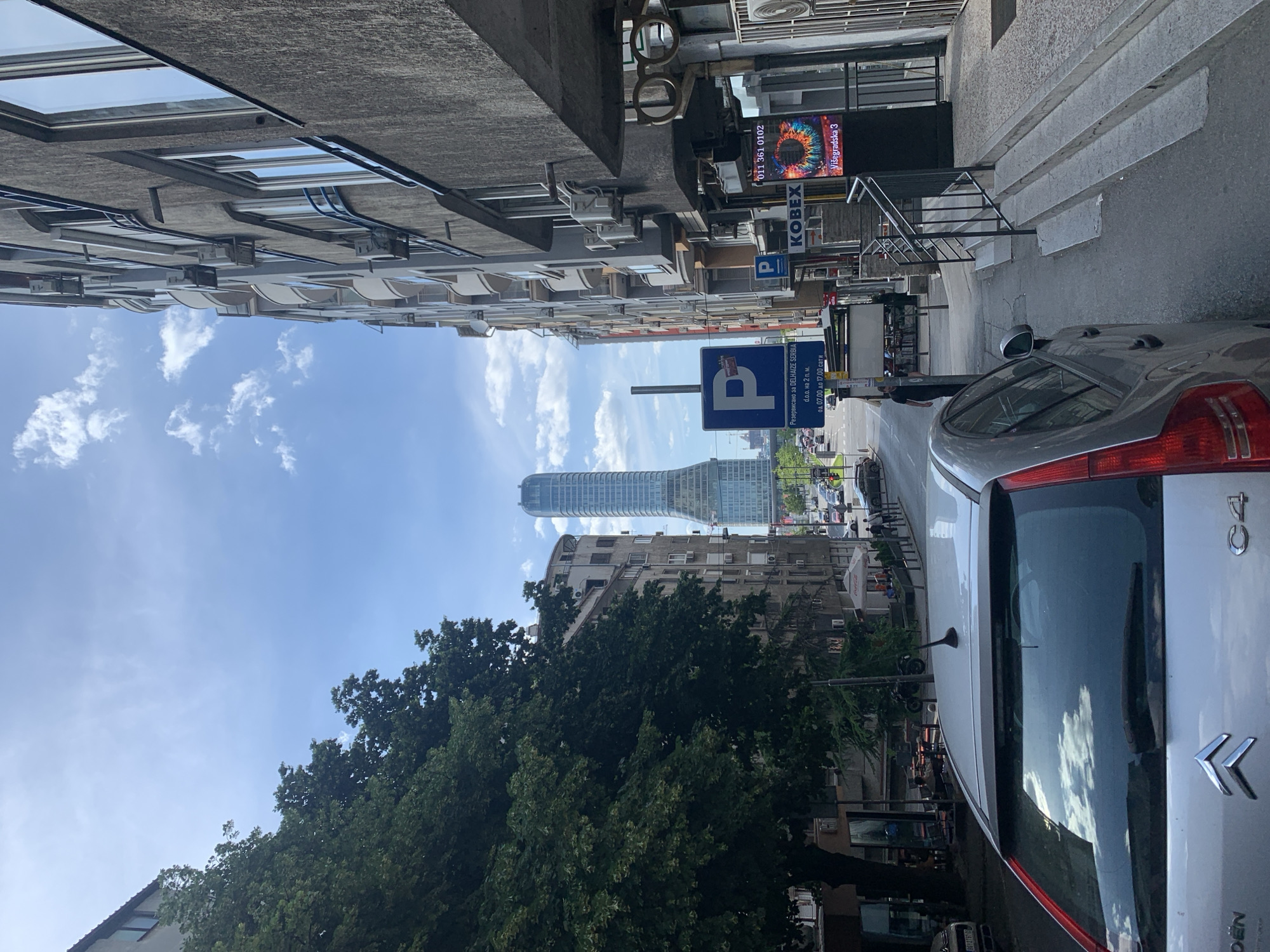 The image depicts a street in Belgrade, Serbia, oriented vertically. On the right side of the frame, part of a silver Citroën C4 car is visible. A parking sign displaying the letter "P" and text related to Delhaize Serbia is prominently featured. Further down the street, a modern skyscraper stands tall amidst older buildings and lush green trees. Buildings line both sides of the street, showcasing a mix of architectural styles. Signage for "KOBEX" and "Višegradska 3" is visible above a storefront. A person is walking along the sidewalk. The sky is blue with scattered clouds.