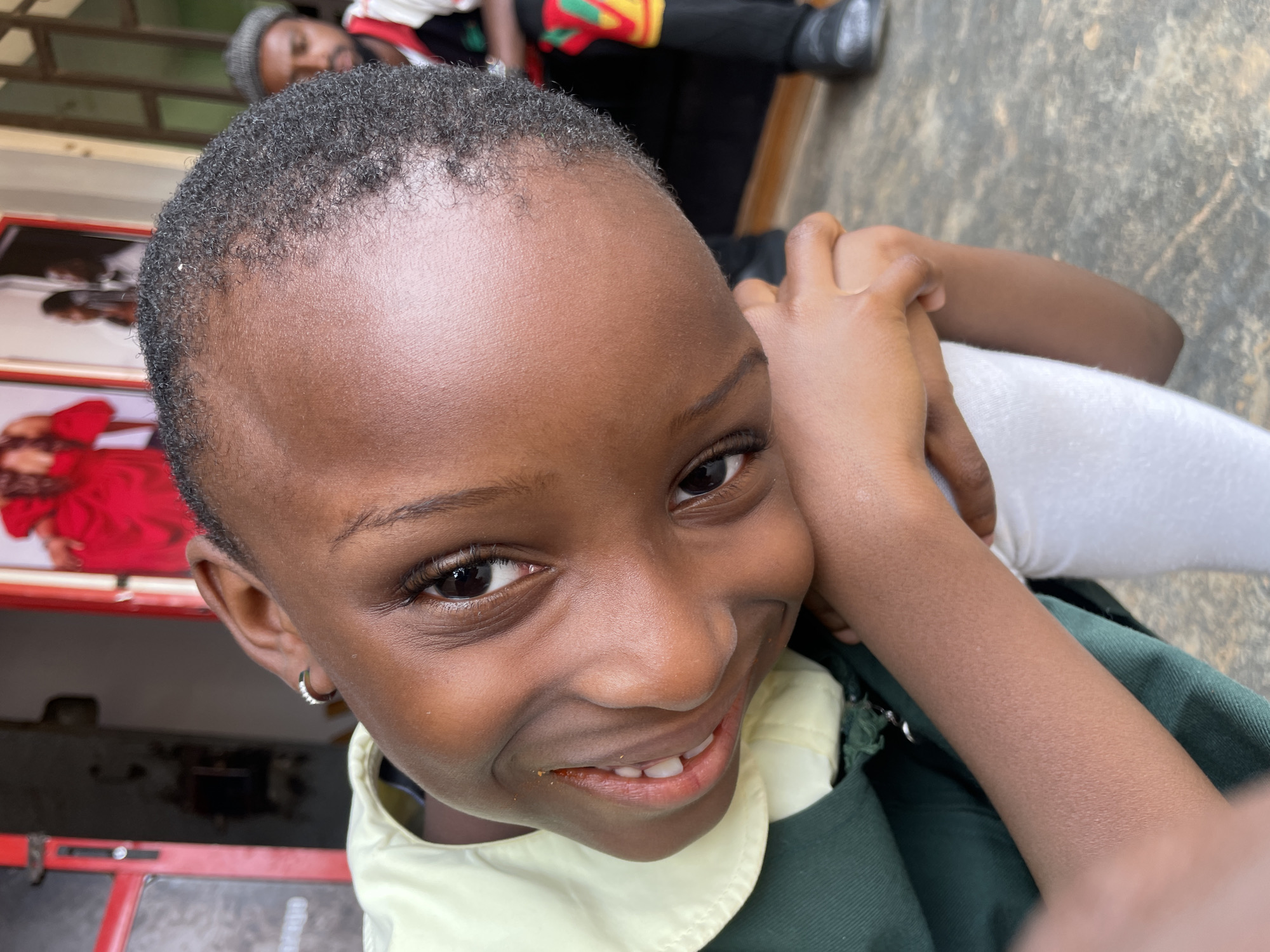 A close-up shot depicts a young, dark-skinned child smiling directly at the camera. The child has short, close-cropped hair and wears a small, round earring. The child is dressed in a dark green uniform with a light yellow collar. In the background, blurred, is a person with dreadlocks and a man wearing black clothing with green, yellow and red stripes. The image appears to be taken outdoors in the Barakin Akawo, Nigeria, due to the environment visible in the background. There are red and black objects in the blurred background along with part of a concrete flooring visible in the top right corner.