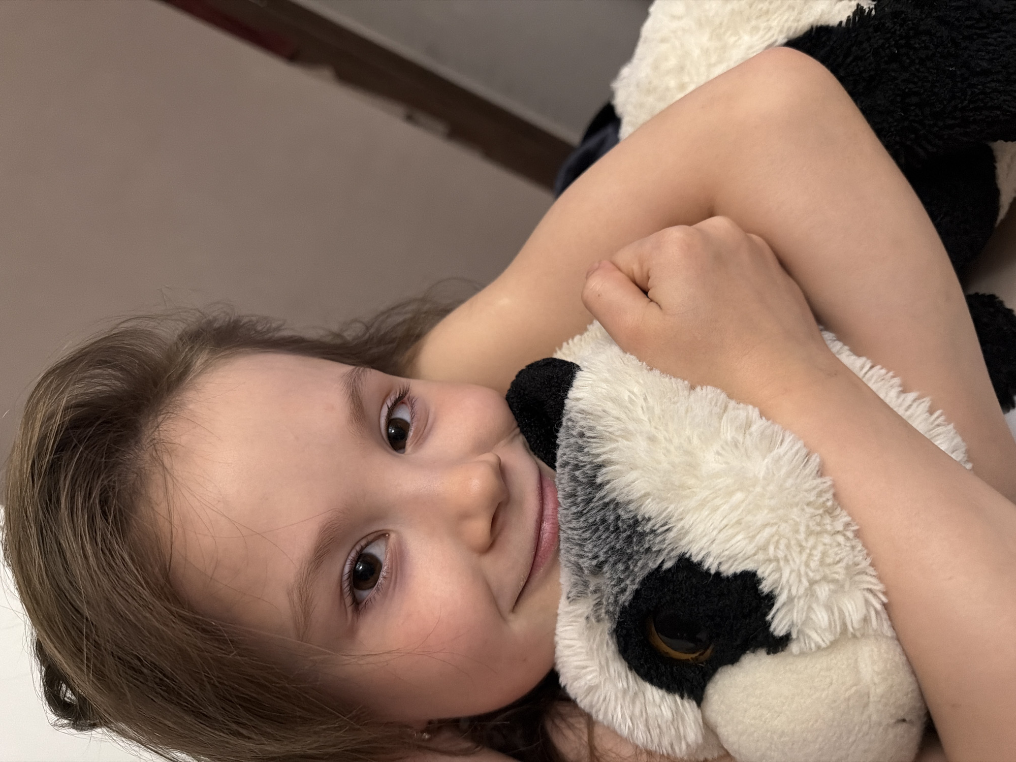 A close-up portrait features a young child, likely a girl, lying down and gently embracing a black and white plush toy. The child has fair skin, light brown hair, and dark, expressive eyes, looking directly at the viewer with a soft, contented smile. Her arms are wrapped around the fluffy toy, which has prominent black and white sections and brown eyes, resembling a panda. The setting appears to be indoors, with a plain, light brown or beige wall forming the background. A darker, horizontal structure is visible in the upper part of the image. The lighting is soft and even, highlighting the child's serene expression and the texture of the toy. The image conveys a sense of warmth, innocence, and comfort. There is no visible text within the frame. While the location is provided as Perm, Russia, the image itself does not contain specific visual cues to indicate this geographical context.