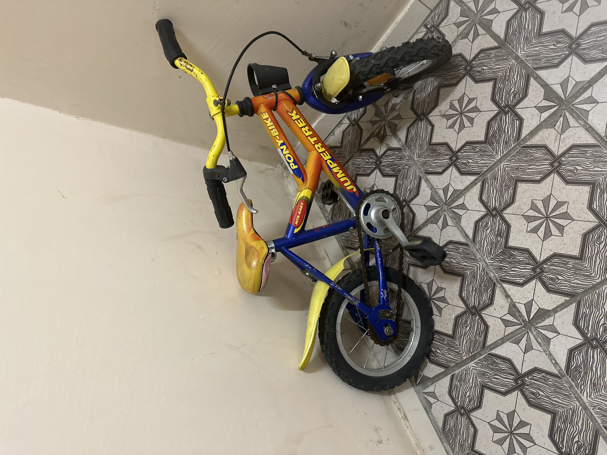A blue and orange JumperTrek Pony-Bike, designed for a very young child, is leaning against the interior corner of a room. The bicycle has a yellow seat and handlebars with black grips. The front wheel is blue with a yellow fender, and the rear wheel is silver with a yellow fender and chain. A small black basket is attached to the handlebars. The bicycle is positioned on a tiled floor with a patterned design of wood grain and geometric shapes. The walls are painted a light, neutral color. No people are present in the scene. The image was taken in Kyiv, Ukraine.
