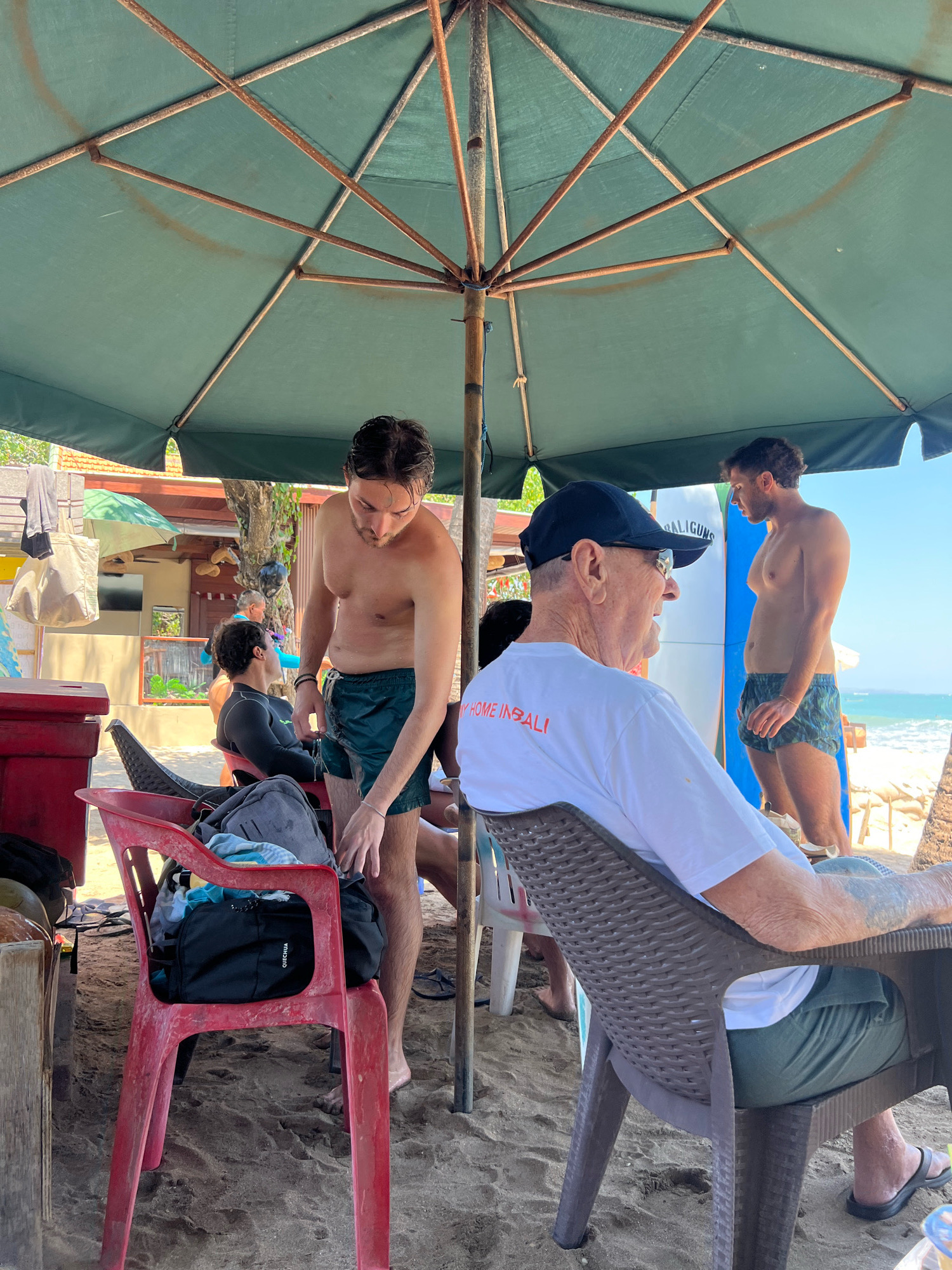 The image depicts a group of people relaxing under a large, green umbrella on a sandy beach, likely in Kuta, Indonesia, given the text on one of the surfboards and the general beach scene. In the foreground, an elderly man wearing a white t-shirt and a black baseball cap is seated in a brown, plastic chair, facing away from the camera. He appears to be engaged in conversation with someone out of frame. To his right, a young man in swim trunks stands looking out towards the ocean. Behind him, another man in dark swim trunks and a black wetsuit top is seated on a red plastic chair. A red cooler is visible to the far left, and various bags and towels are scattered around the seating area. Surfboards stand upright near the young man in swim trunks. The environment is sunny with a clear blue sky, and the ocean is visible in the background, with gentle waves. The sand is light brown and appears somewhat damp.