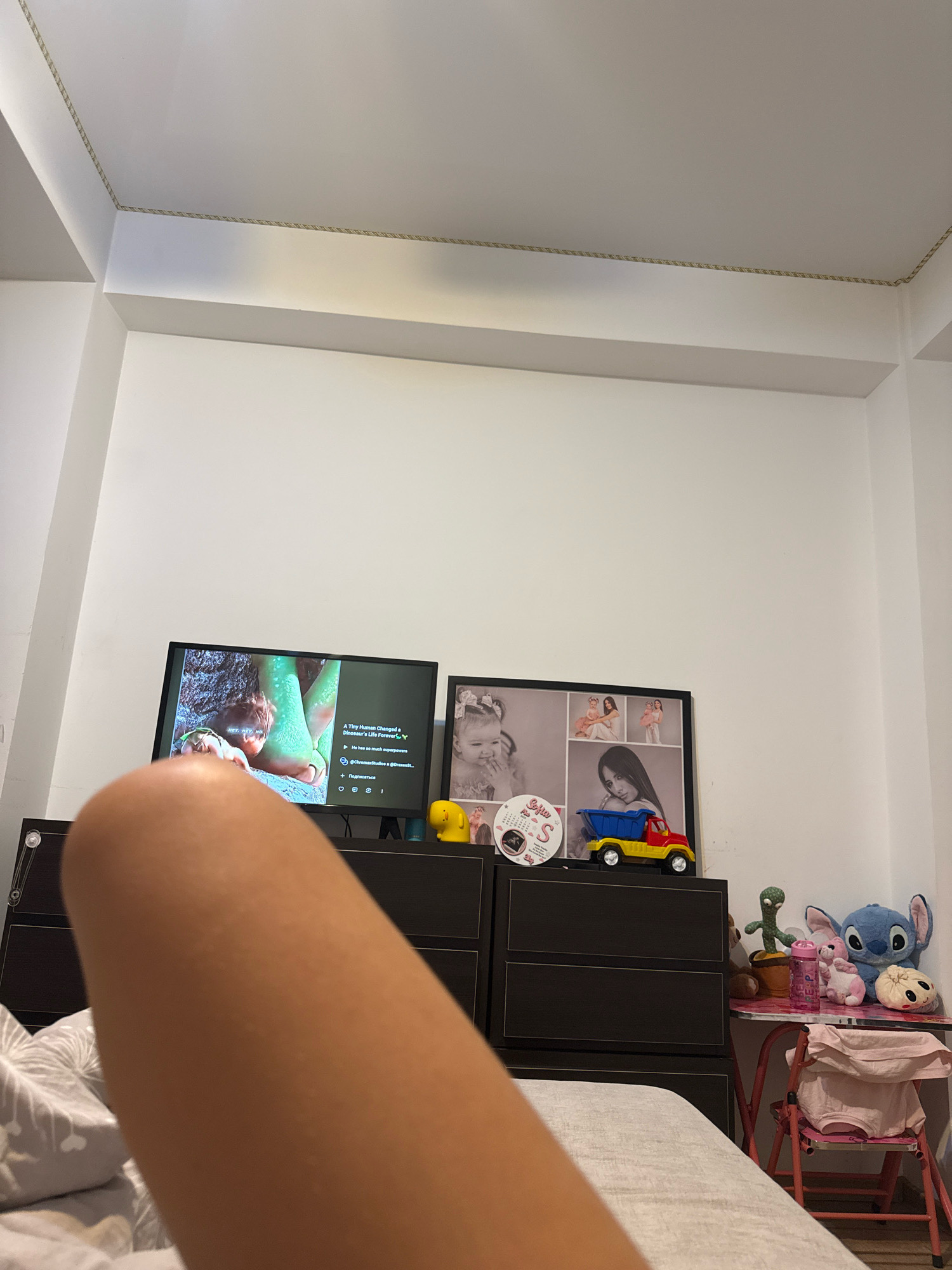 In a bedroom, a television displays a video of a child with a dinosaur. To the right of the television, a framed photo collage features images of a baby and a young girl. A yellow duck figurine and a toy dump truck sit atop a dark dresser. On a small pink table, a plush cactus, a stuffed bunny, a water bottle, and a Stitch toy are arranged. A pink chair with a light pink garment draped over it is situated to the right of the table. The room has white walls and a white ceiling with decorative trim. The image is taken from a low angle, with a person's leg in the foreground. There is no information to suggest the location is Chișinău, Moldova.