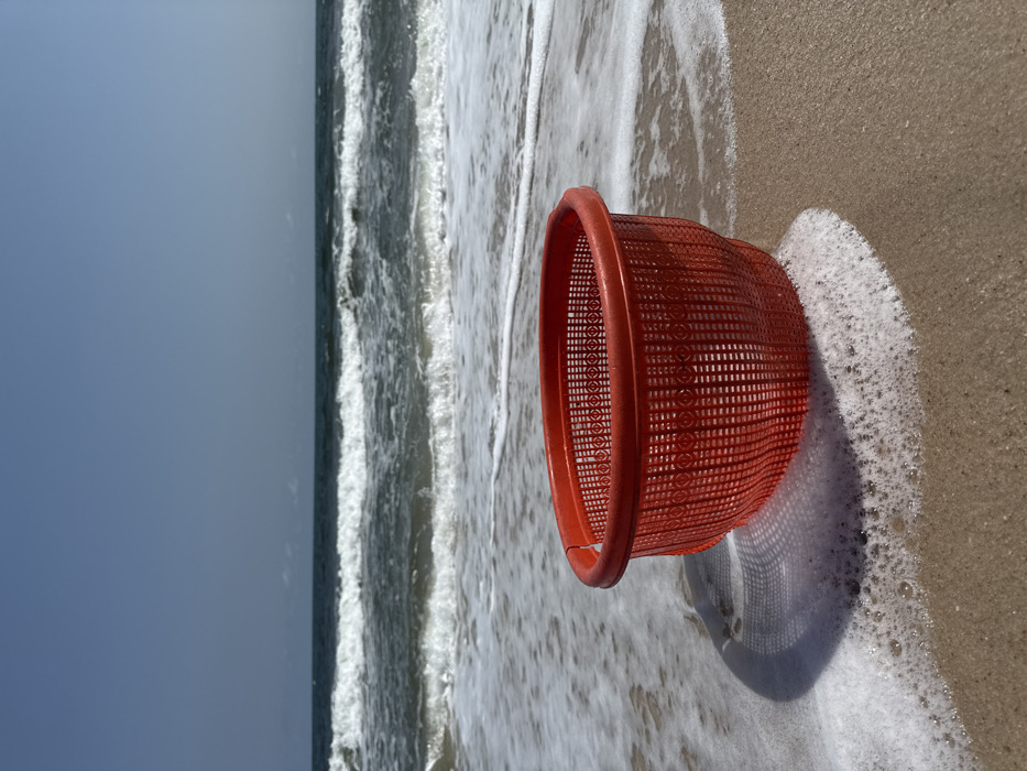A red, circular plastic basket with a perforated diamond and circle pattern is positioned on a light brown sandy beach in Mopo Onijebu, Nigeria. The basket occupies the lower center of the frame. White seafoam from an ocean wave surrounds the left side of the basket's base, partially obscuring its shadow on the sand. In the midground, multiple lines of breaking ocean waves with white crests extend across the dark blue-green water. The background consists of a clear, pale blue sky meeting the ocean horizon. No individuals are present in the scene. The overall lighting suggests a clear, sunny day.