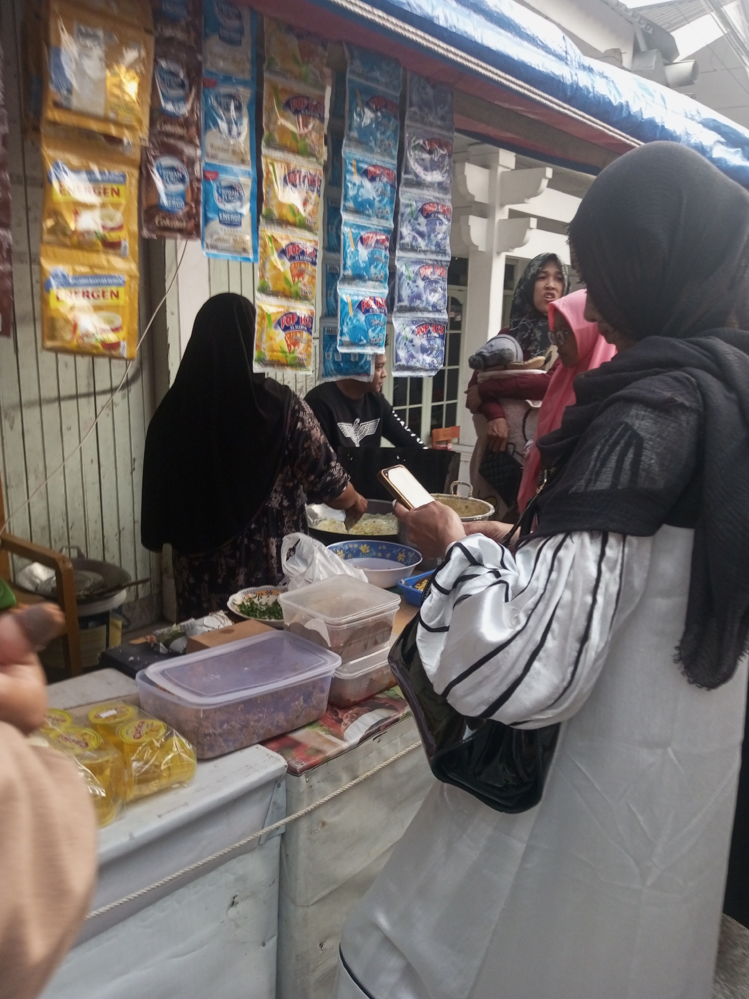 A street food stall is pictured in Pandansari, Indonesia. A vendor in a black hijab is preparing food. To her right, another woman in a pink hijab and a person in a black hoodie are visible. The stall displays various packaged food items hanging from above, including what appear to be instant noodle and beverage sachets. On the counter, there are containers of food ingredients, plastic bags of small yellow-packaged items, and a mobile phone in the vendor's hand. The stall is set up against a wooden-paneled wall. The scene suggests a typical daily operation of a street food vendor in the area.