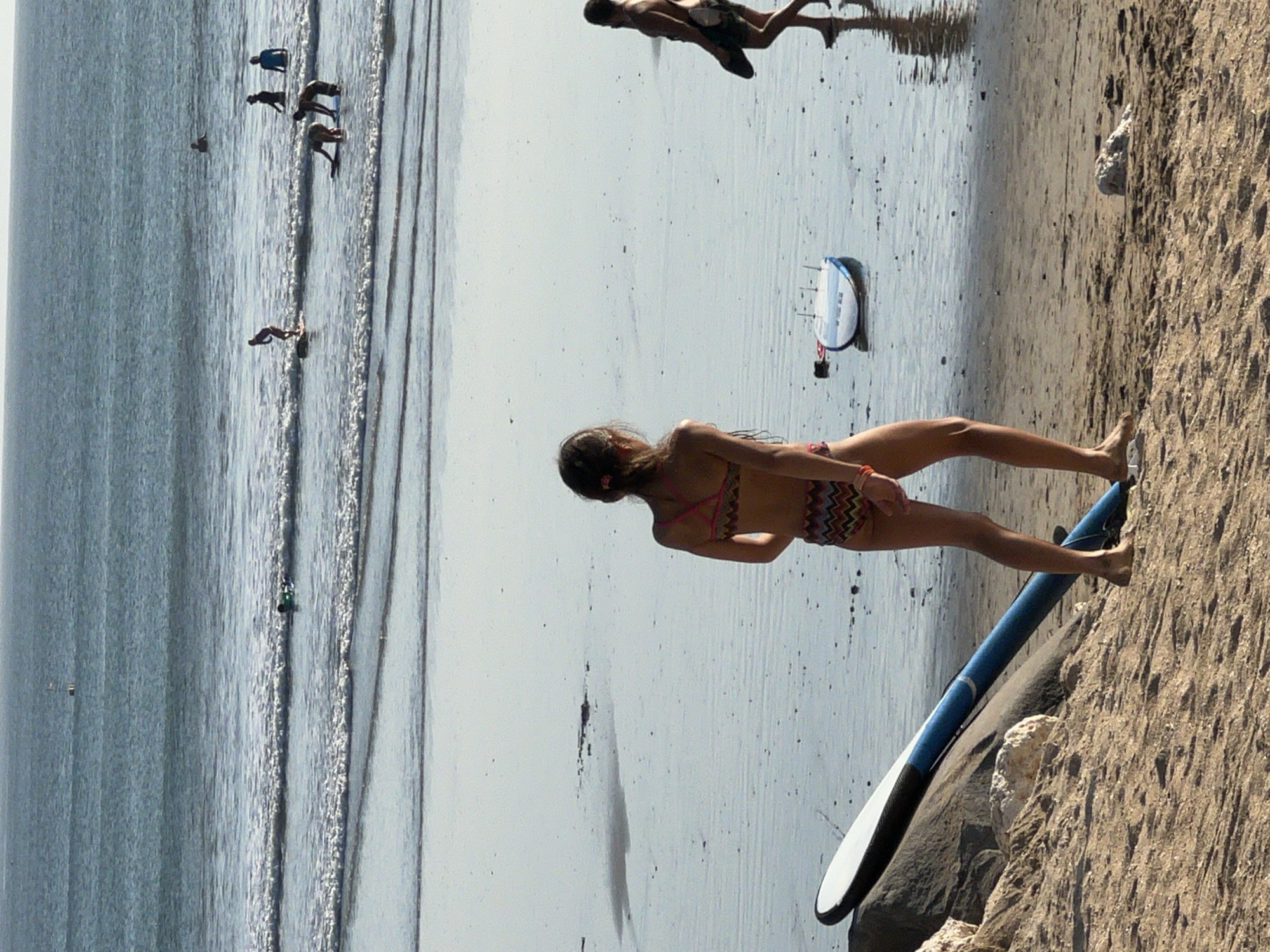 The image shows a beach scene at Kuta, Indonesia. Several people are visible, some in the water and some on the sand. In the water, some individuals appear to be surfing or bodyboarding. One person is lying prone on a surfboard in the waves, while others are further out in the ocean. On the shore, a woman in a bikini is standing with a paddleboard. The water is calm, with small waves rolling in. The sand is light brown and appears wet near the water's edge. The sky is bright, suggesting it is daytime, likely midday or afternoon. There is a clear division between the water and the sandy beach. The overall atmosphere seems relaxed and recreational, with people enjoying water activities.