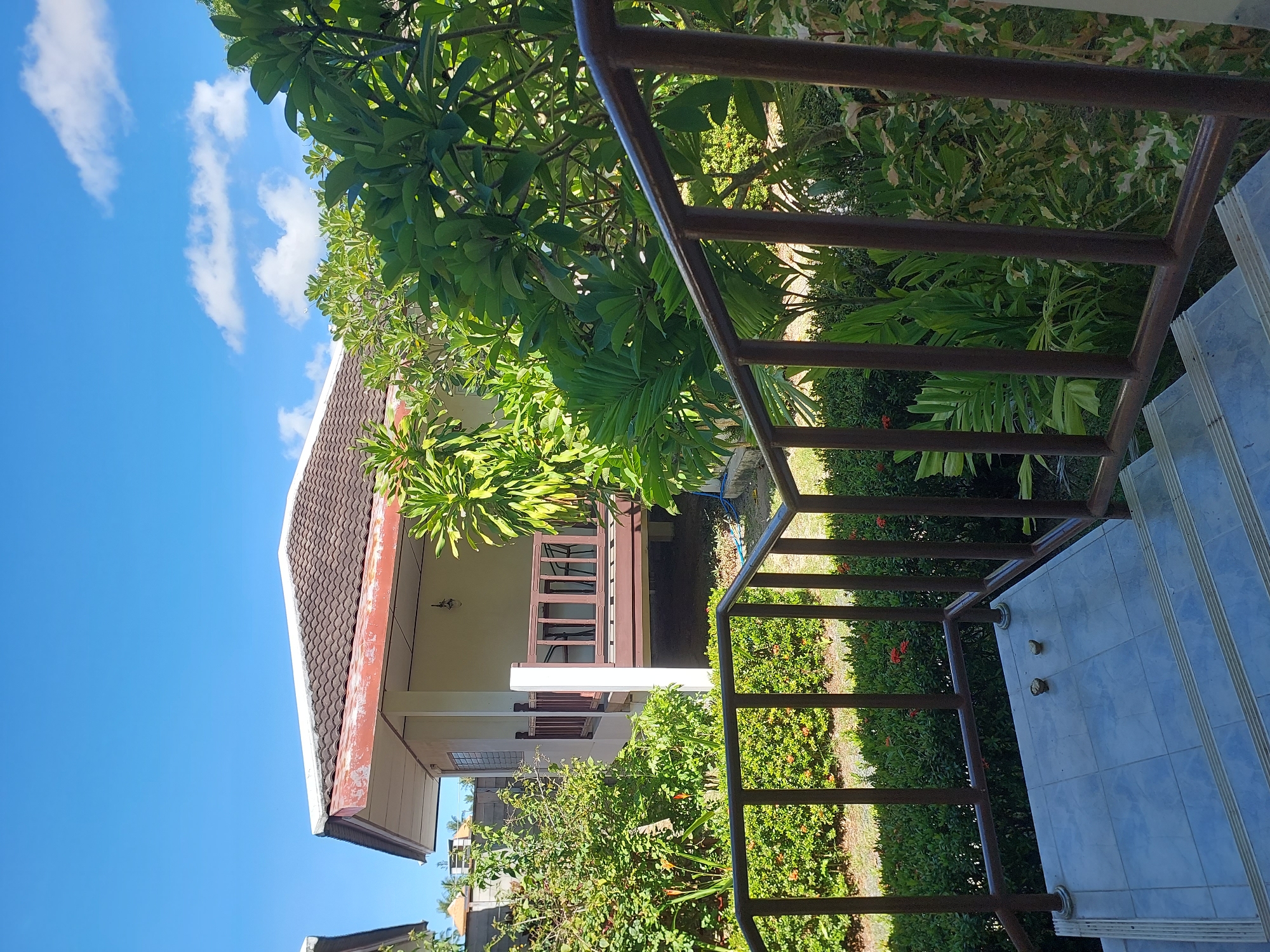 The image shows a view from a tiled staircase with a brown metal railing. The staircase leads up to a small, light-colored building with a brown tile roof. The building has a covered porch with a wooden railing. Lush green foliage, including palm trees and other tropical plants, surrounds the building. The sky is blue with scattered white clouds. The scene appears to be in a tropical setting, consistent with Baan Mae Nam, Thailand.