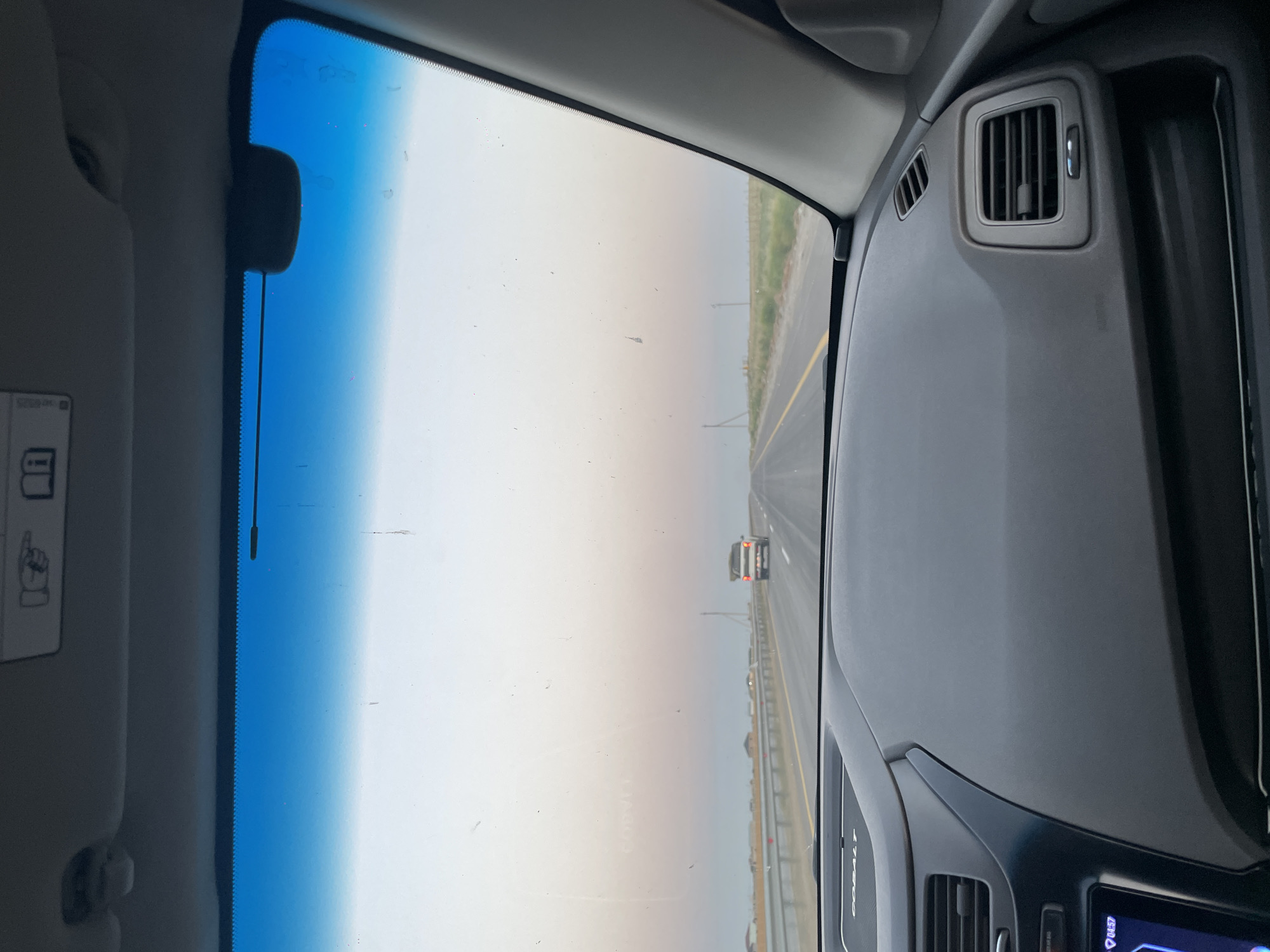 The image is taken from inside a car, looking out through the windshield. The dashboard of the car is visible in the foreground. The road ahead is straight and appears to be a highway. There is a white pickup truck driving in the same direction as the camera car. The sky is a pale gradient of blue and pink, suggesting it might be dawn or dusk. The environment appears to be rural or on the outskirts of a town, with sparse vegetation and a flat landscape. There are no people clearly visible in the image besides the driver implied by the camera's perspective. The car appears to be in motion, driving on a paved road. The location is specified as Toksanbay, Kazakhstan.