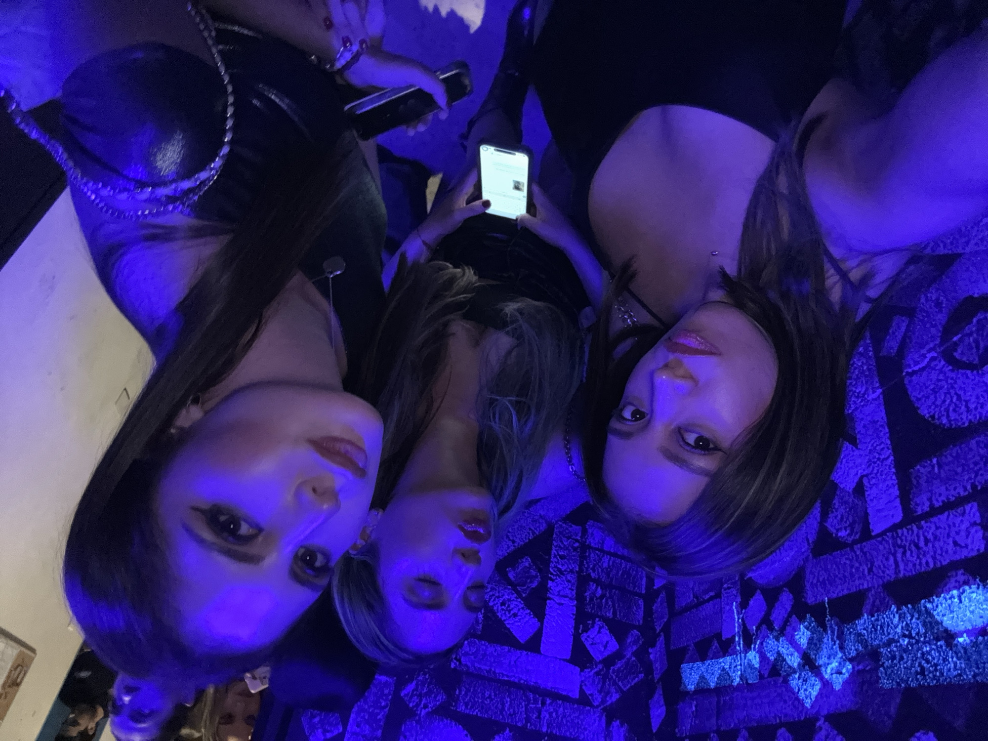 Three women are posing closely for a photo, all angled towards the camera. The woman on the left is wearing a dark, shiny top with a decorative chain strap. The woman in the center is holding and looking at a smartphone. The woman on the right is wearing a low-cut top with a necklace. The background is a dark surface with a geometric pattern lit with a blue light. It is impossible to determine from this image if this photo was taken in San Bernardino Tlaxcalancingo, Mexico.