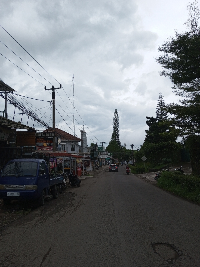 The scene depicts a street in Melakudikadu, India, under an overcast sky. The two-lane asphalt road extends into the distance, showing some surface wear and a small pothole in the mid-right foreground. On the left, a dark blue pickup truck is parked facing the viewer, displaying license plate "B 9031 DDB". Behind the truck, a commercial area features buildings, one with a sign reading "HANDY'S ALUMINIUM STAINLESS STEEL," and several open-fronted stalls with awnings. A utility pole heavily laden with multiple overhead electrical and communication wires stands centrally on the left. Further down, a tall, slender communications tower and a building with a spire-like structure are visible. On the right side of the road, dense green foliage and several tall, narrow coniferous trees line the street, with a white road sign partially visible among them. Two vehicles are in motion on the road, moving away from the viewer: a dark-colored sedan followed by a person on a red motorcycle. Several other motorcycles are parked along the left roadside near the stalls and along the right roadside among the greenery. No pedestrians are clearly visible.
