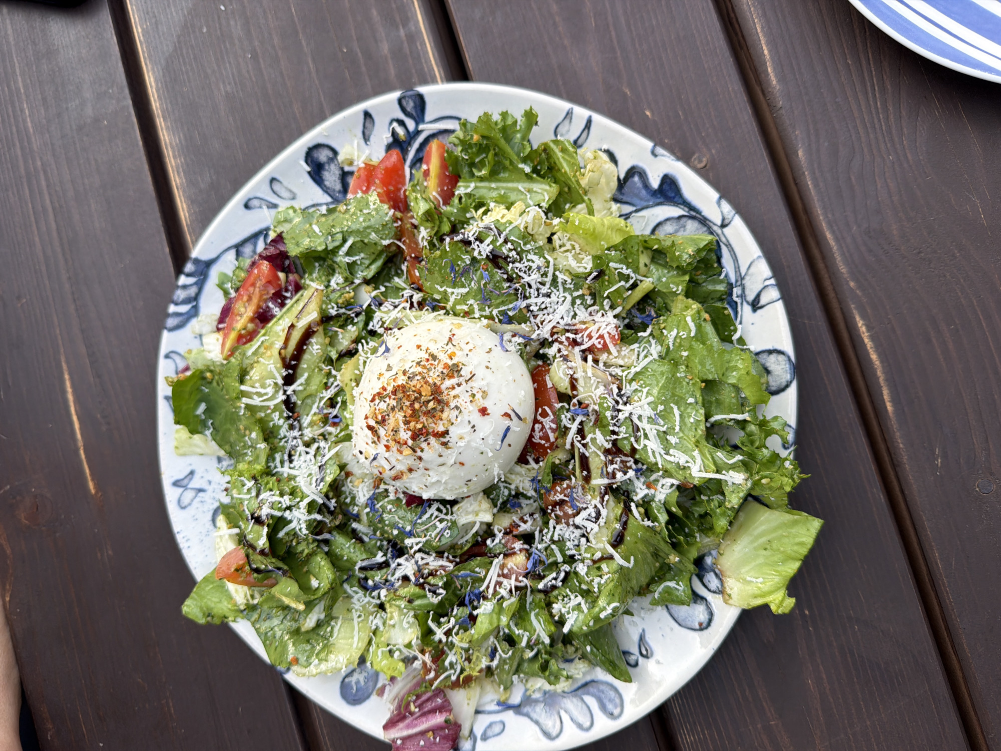 A close-up, overhead shot shows a salad on a patterned white plate with blue designs. The salad is a mix of various leafy greens, including romaine lettuce, radicchio, and possibly kale. It is topped with halved cherry tomatoes, shredded cheese, and a large ball of burrata cheese in the center. The burrata is sprinkled with red pepper flakes and other seasonings. Small blue flower petals are scattered throughout the salad, and a dark drizzle, likely balsamic glaze, coats some of the ingredients. The plate rests on a dark brown wooden table with a visible grain. The setting appears to be outdoors, suggesting a restaurant patio or garden. There are no people visible in the frame. Ventspils, Latvia is not discernible from the image.