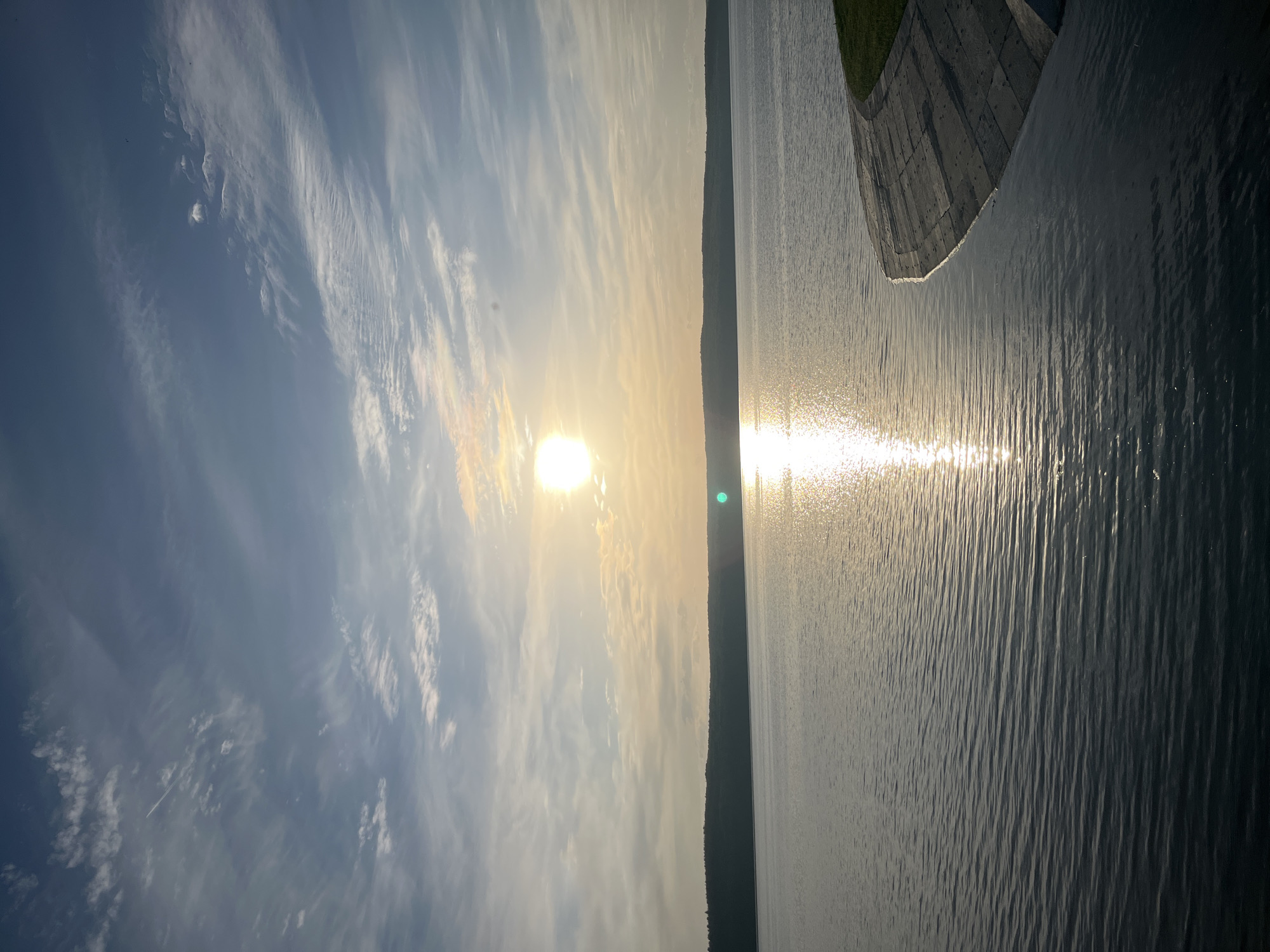 The image depicts a body of water, likely a lake or river, reflecting the sunlight. A forested shoreline is visible in the distance. The sky above is filled with clouds. In the upper right corner, there appears to be a concrete embankment. The location is presumably in Chaikovsky, Russia. No people or specific actions are visible in the image.