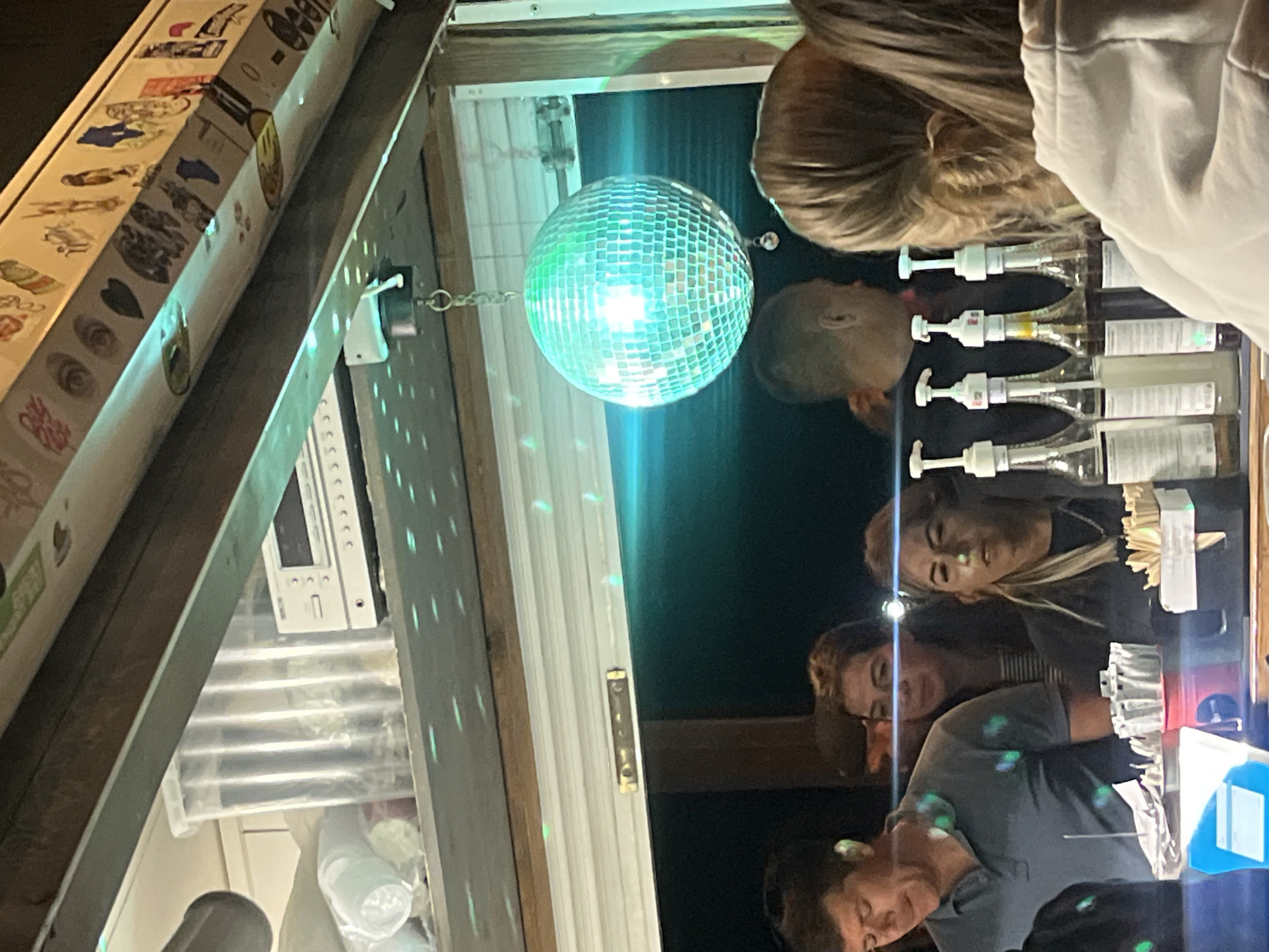 This is a medium shot, taken at night, of people behind a counter, likely a bar or food service establishment, in Pionersky, Russia. In the background, a disco ball hangs from the ceiling, reflecting green light onto the surfaces. On a shelf above the counter, a clear plastic container with items inside and a white electronic device are visible. Several people are visible behind the counter. A man in a blue polo shirt is on the left. A woman with dark hair and stripes on her shirt is behind him. To her right, a woman with blonde hair is looking forward. In front of them, four bottles with pumps, likely containing syrups or sauces, are arranged in a row. The scene is dimly lit, with the disco ball's light providing the primary illumination.